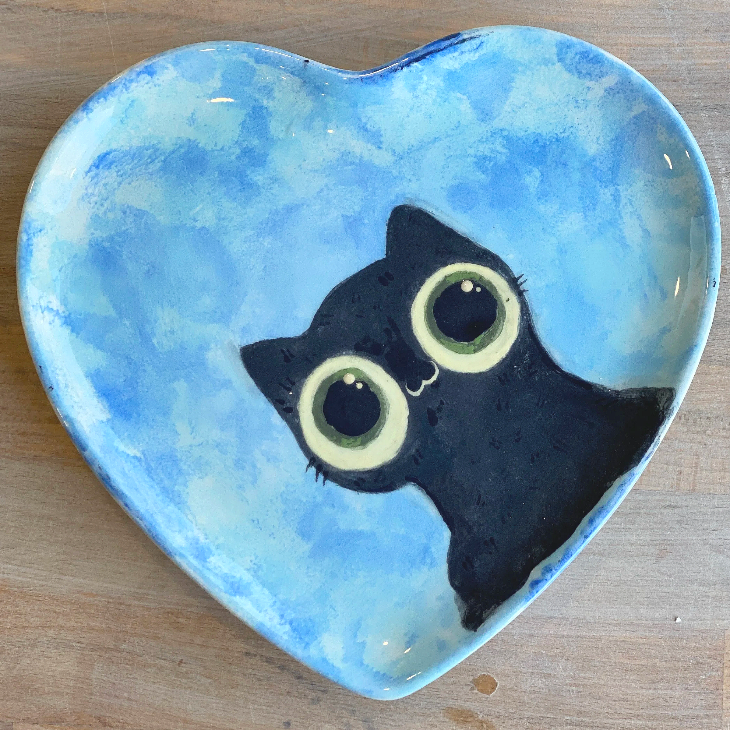 Heart-shaped ceramic dish with a painted black kitten face and large green eyes, set on a wooden surface.