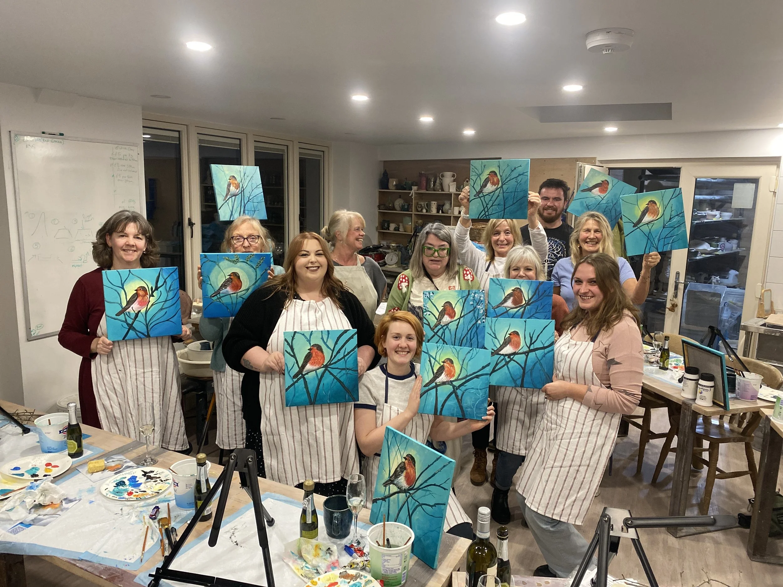 Group of people in a kitchen or art studio holding up completed bird paintings on canvases, smiling and showing their art.