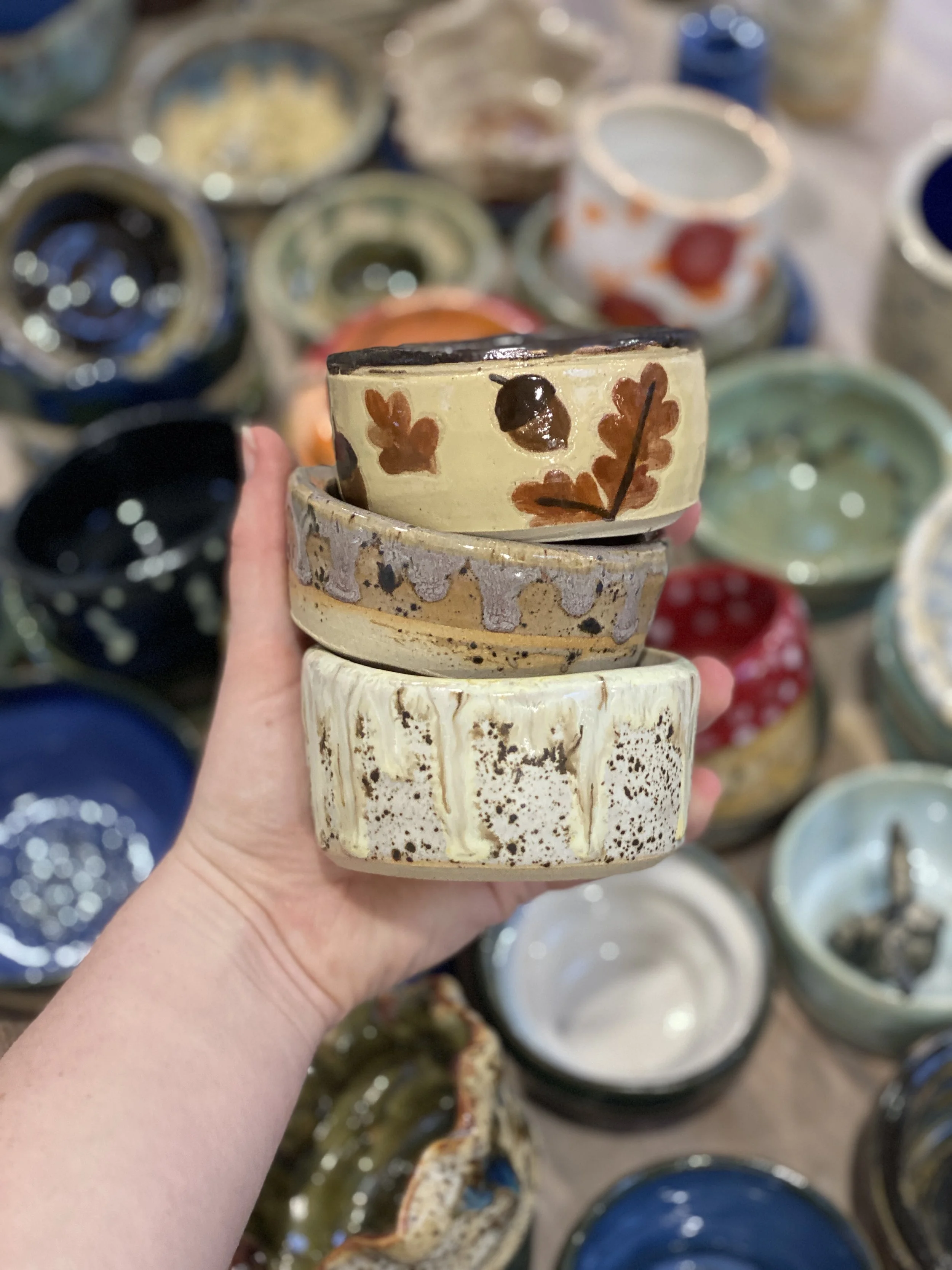 A hand holding three stacked ceramic bowls in front of a display of various colorful bowls and pottery.