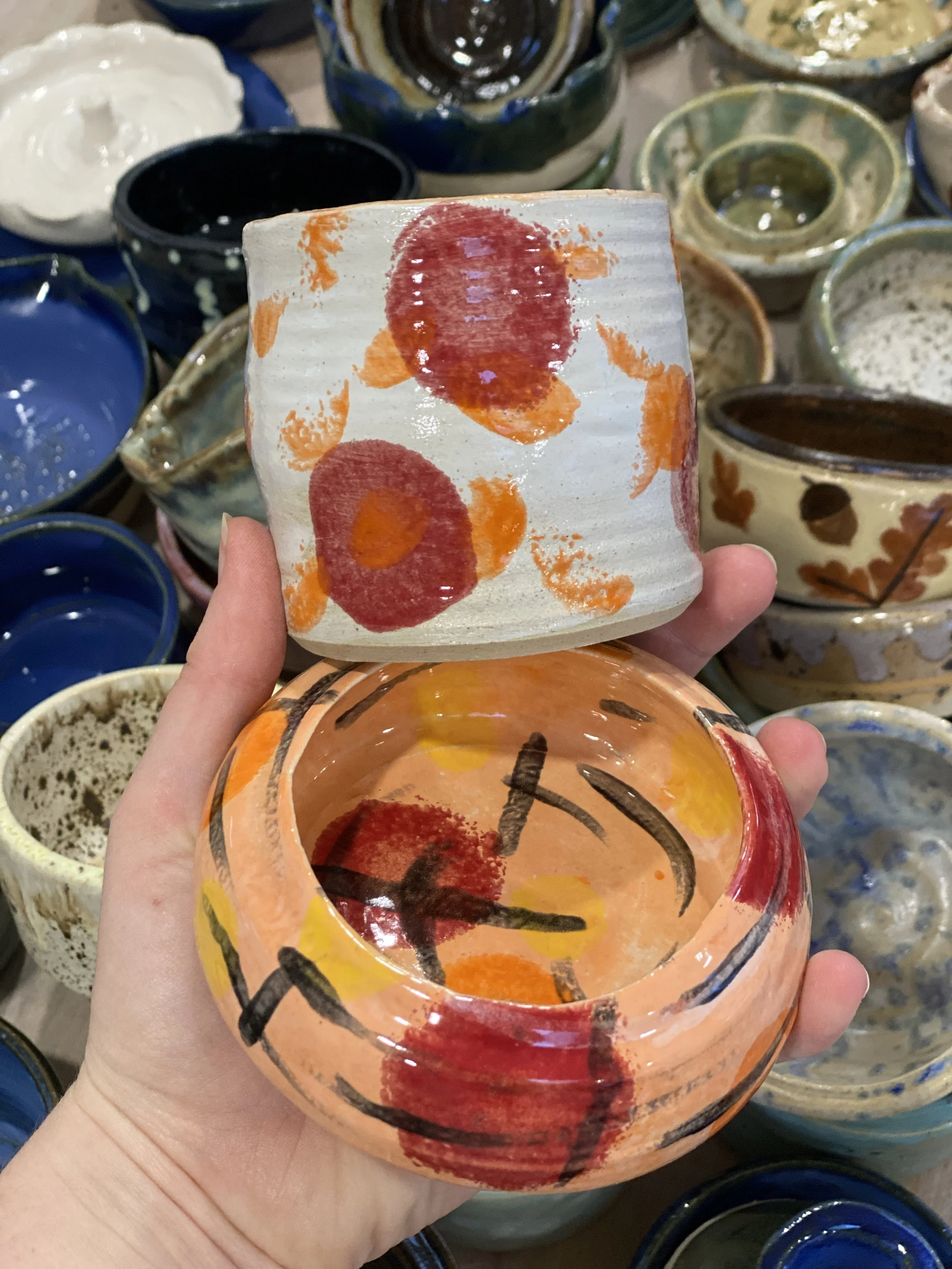 Two hand-painted ceramic bowls, one with a lid, are held up against a background of many other colorful ceramic bowls on display. The bowls feature abstract red, orange, yellow, and black designs.