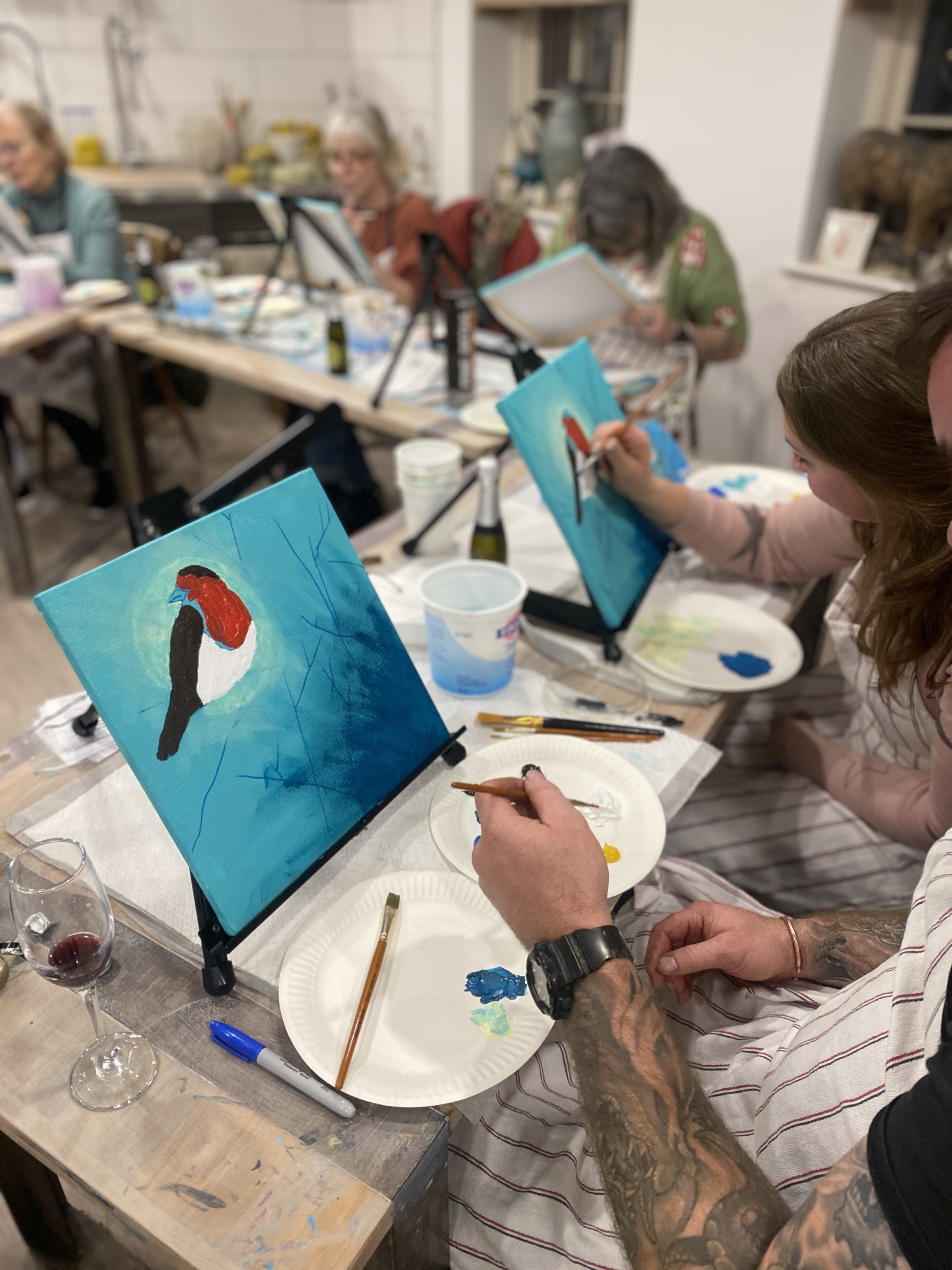 People painting bird pictures on canvases at an art class in a room with art supplies.