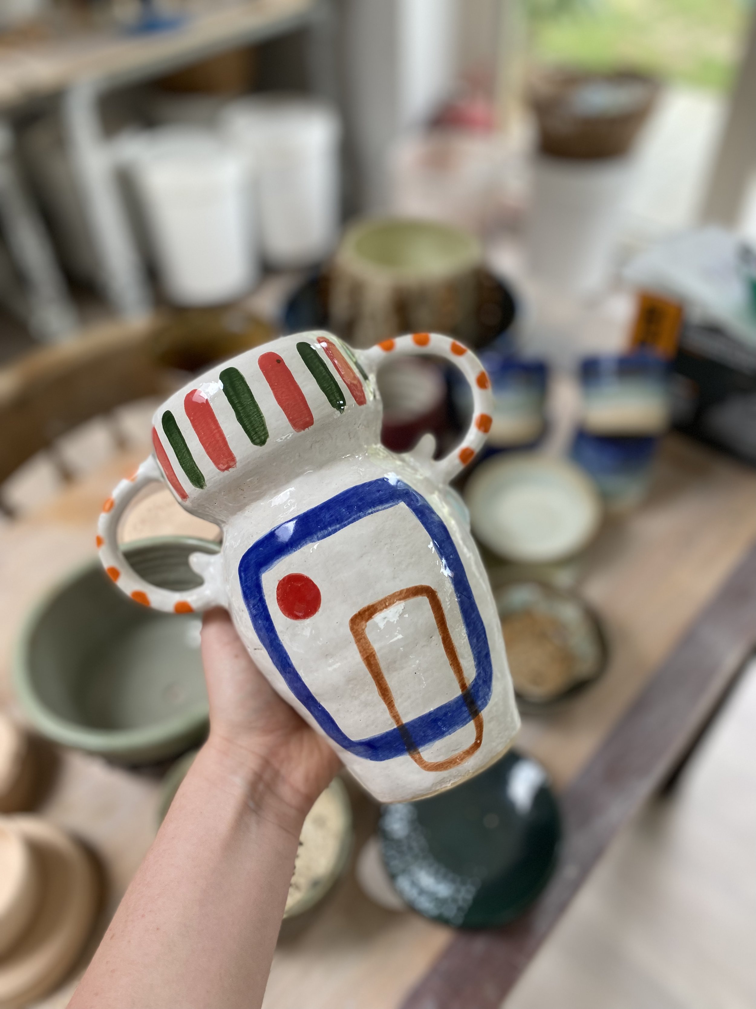 Colorful ceramic pitcher with decorative stripes held in front of a table full of various ceramic bowls and vessels.