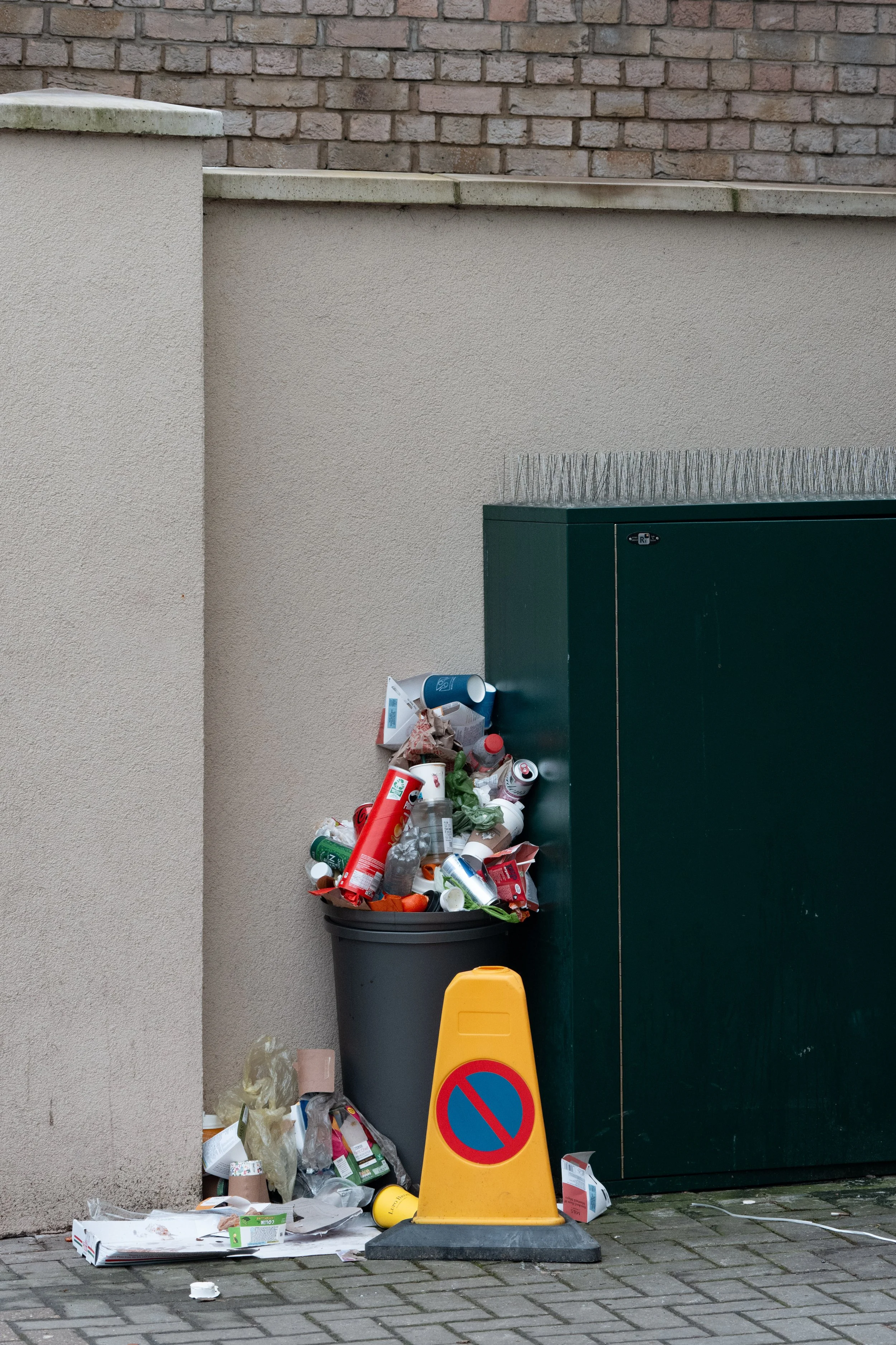 Overflowing trash can with discarded waste and a yellow no parking cone in front of it, against a wall.