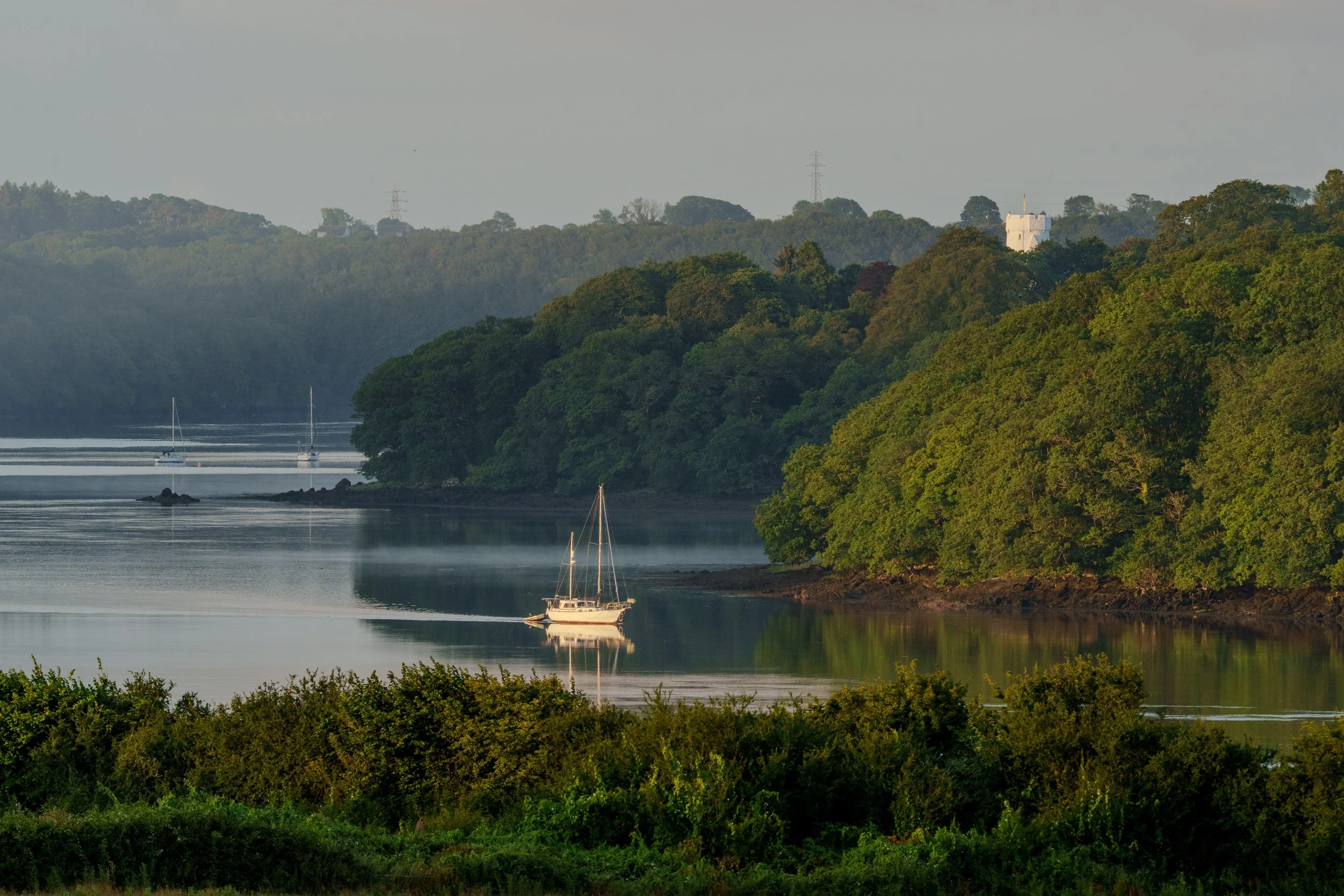 A sailboat on a calm river surrounded by green trees and hills, with a white castle on top of the hill in the background.