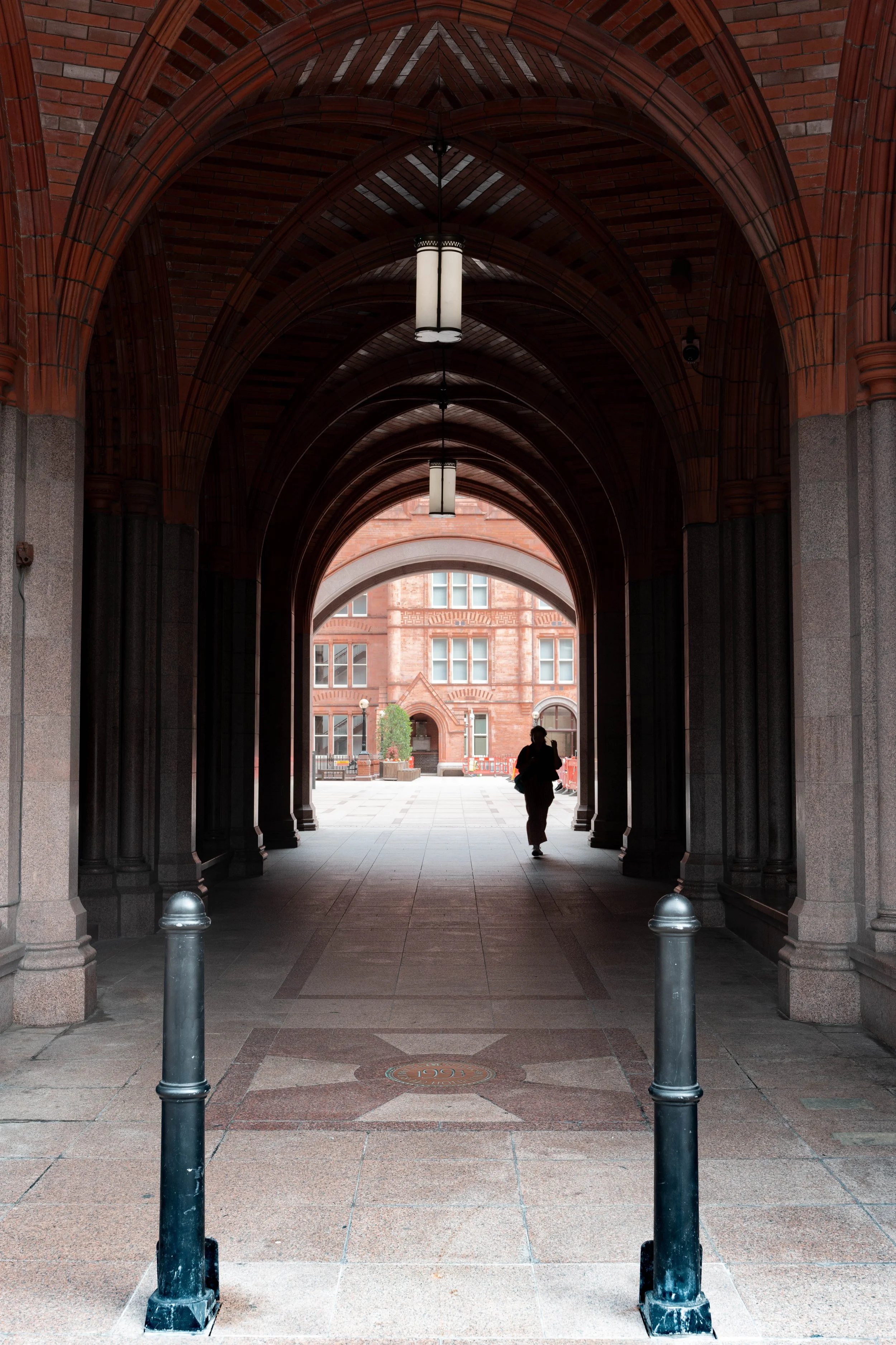 A person walking through an arched brick passageway with hanging lights, leading to a courtyard with a red brick building in the background.