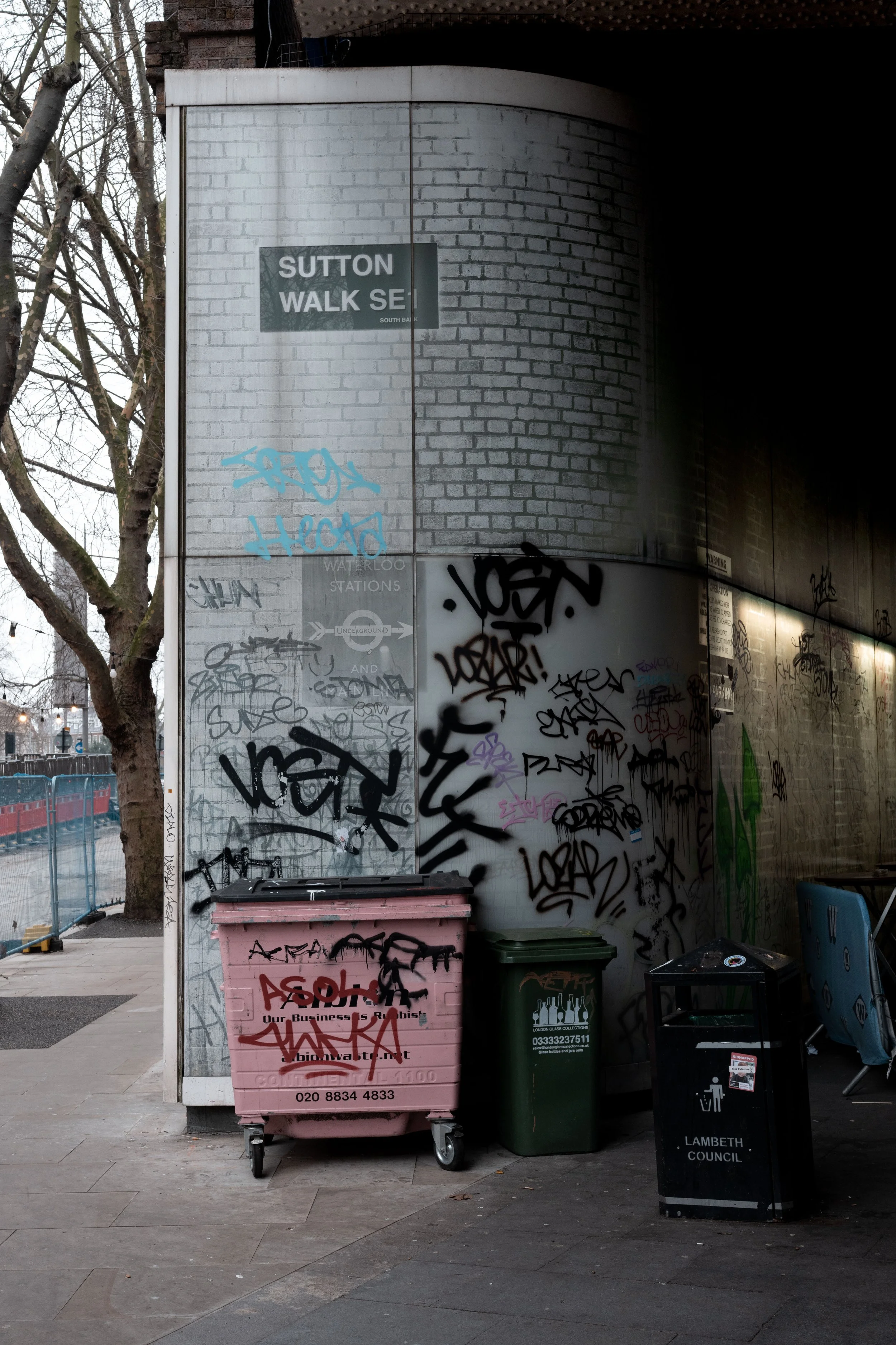 Street scene at a corner with graffiti-covered walls and multiple garbage bins, including a pink bin, a green bin, and a black bin labeled 'Lambeth Council'. A street sign reads 'Sutton Walk SE1'. Empty chairs are nearby, and a tree with leafless bra