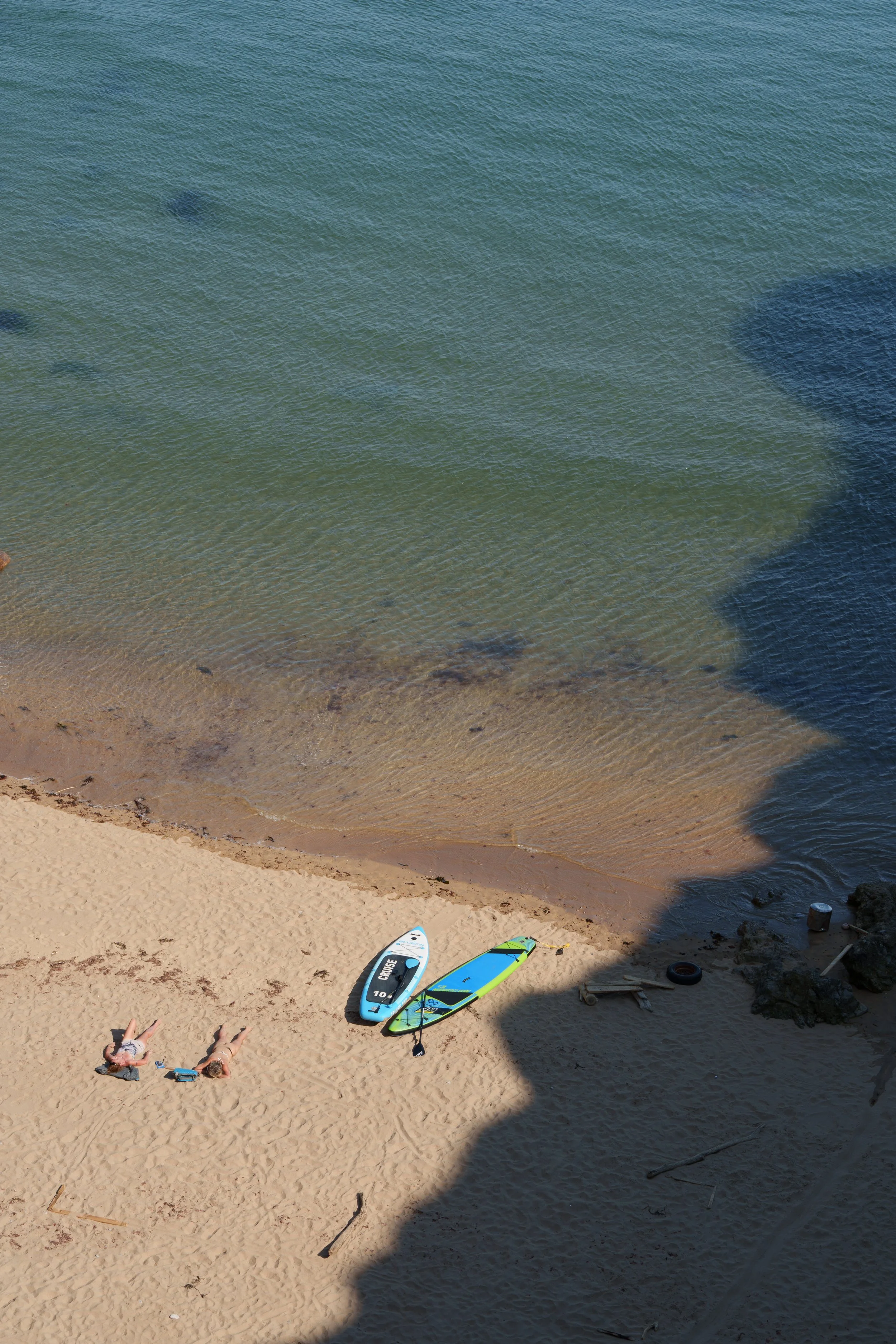 A beach scene with two people lying on the sand, a paddleboard, and a surfboard resting on the sand near the water's edge. The shoreline shows gentle waves lapping against the sand, and there is a shadow cast over part of the beach.