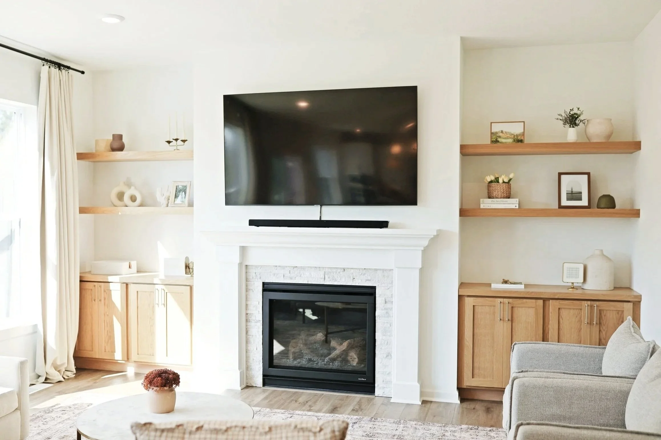 Living room with a white fireplace and a large flat-screen TV mounted above, beige armchair, wooden shelves with decorative items, and cream curtains by the window.