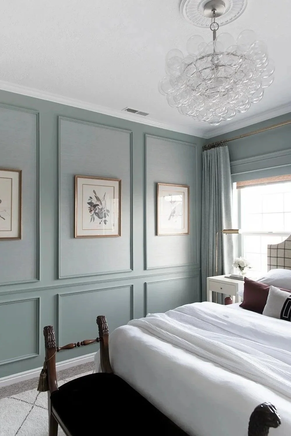 A bedroom with light blue walls, three framed art pieces, a white nightstand with a flower arrangement, a window with curtains, a crystal chandelier, a bed with white bedding and decorative pillows, and a wooden chair at the foot of the bed.