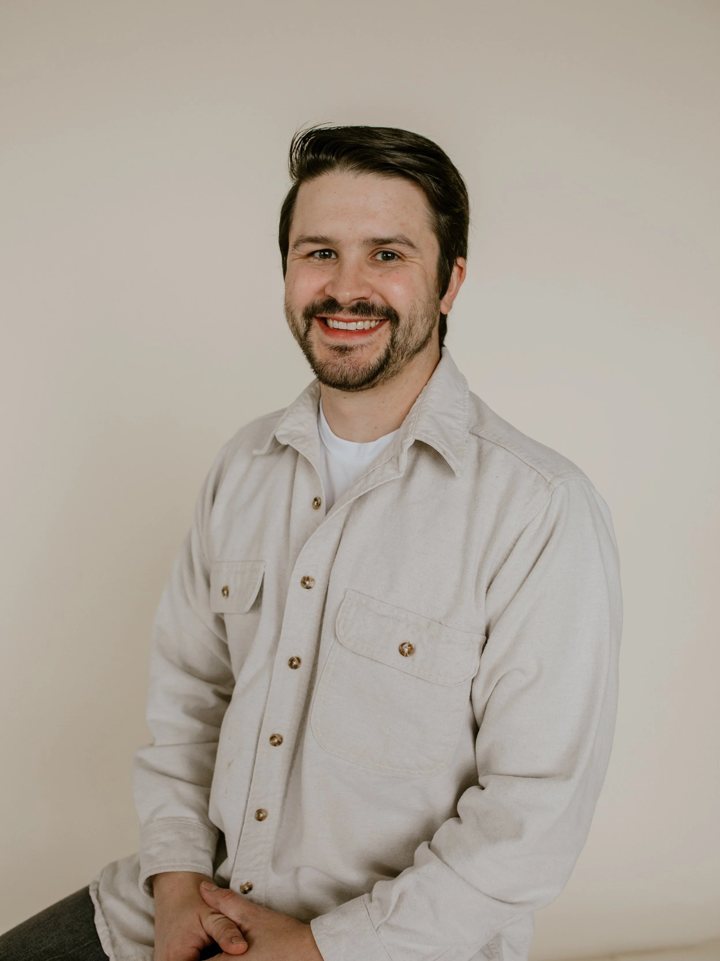 A man with dark hair and a beard, wearing a beige button-up shirt over a white t-shirt, smiling and sitting against a plain, light-colored background.