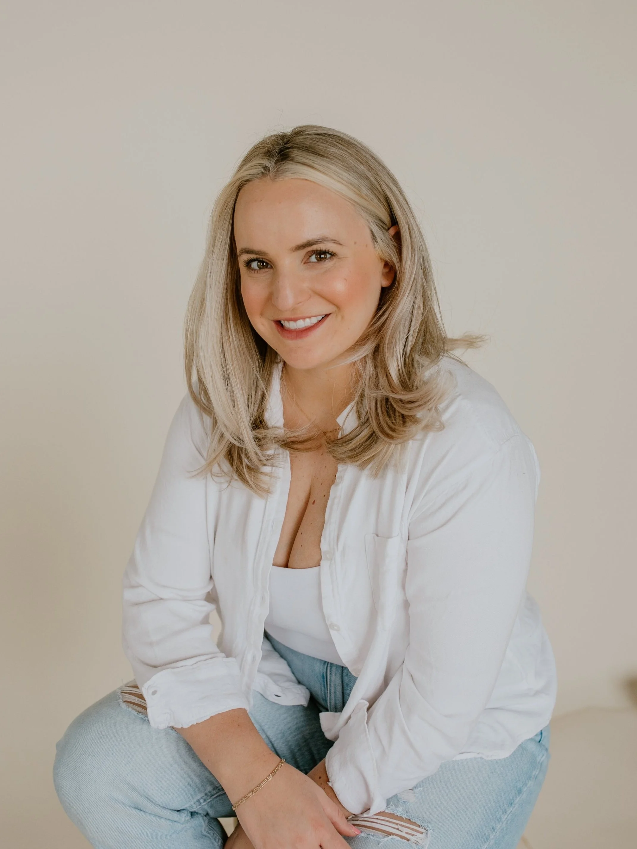 A smiling young woman with blonde hair, wearing a white shirt and ripped jeans, sitting against a plain light background.