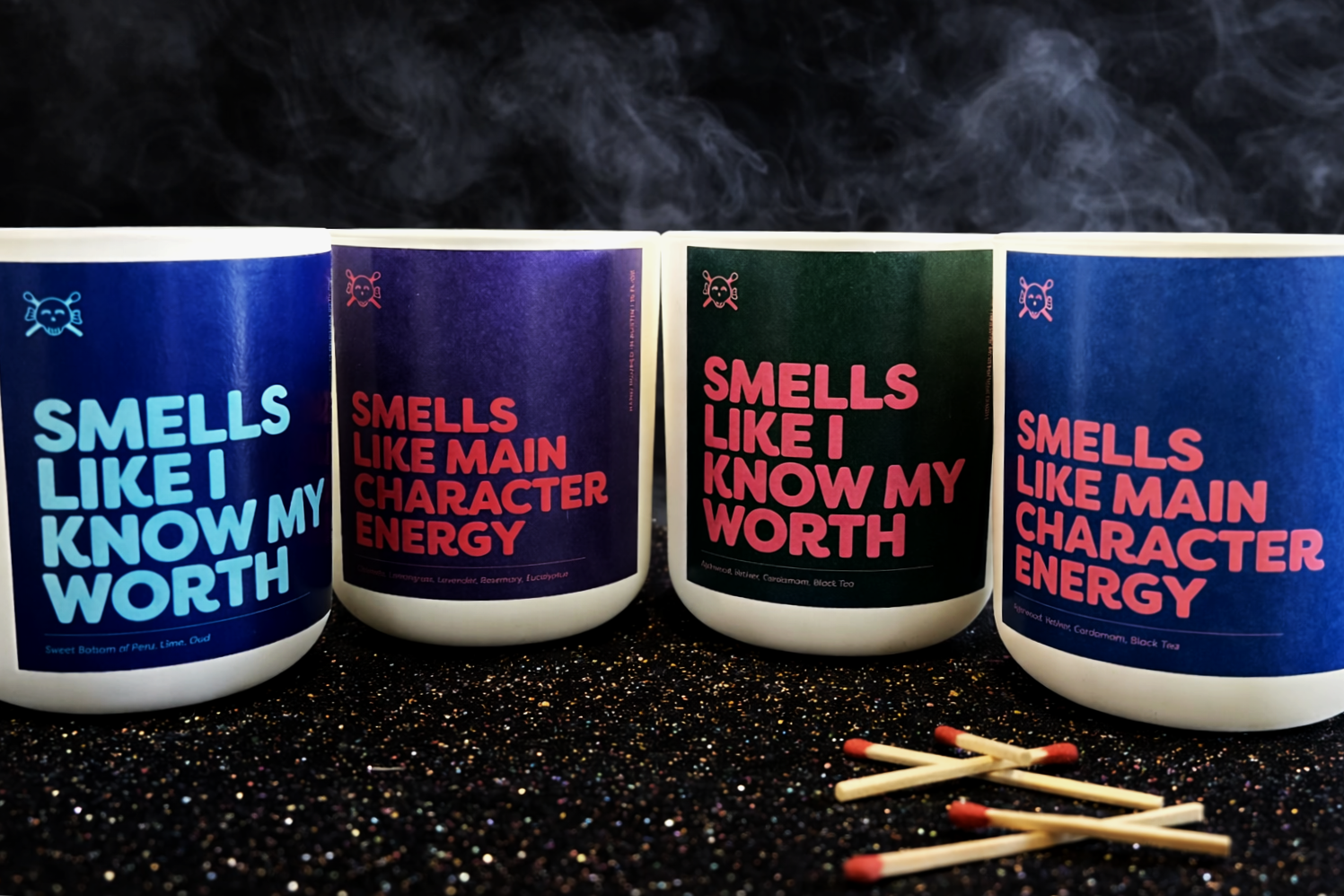 Four cups with colorful labels that read, 'Smells Like I Know My Worth' and 'Smells Like Main Character Energy,' placed on a black glittery surface with matchsticks in front, and smoke rising above.