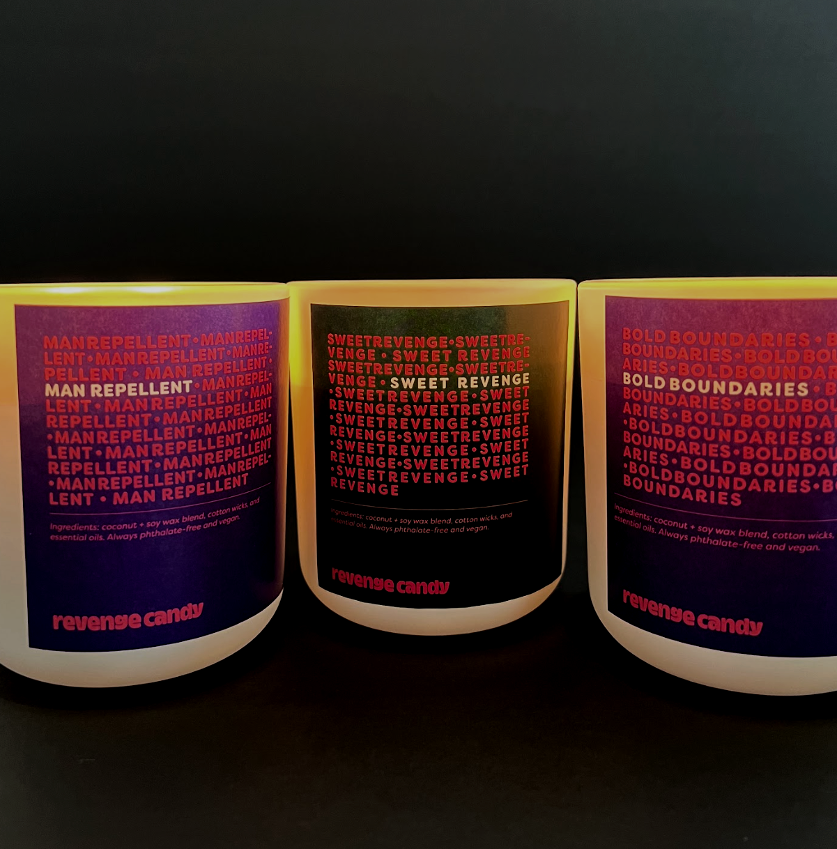 Three candles with dark labels and bright borders, each titled with different words or phrases. The left candle is labeled "MANREPELLENT" with ingredients and description, and has the words "revenge candy" in pink at the bottom. The middle candle is labeled "SWEET REVENGE" with similar ingredients, and also has "revenge candy" at the bottom. The right candle is labeled "BOLD BOUNDARIES" with ingredients and the same bottom phrase.