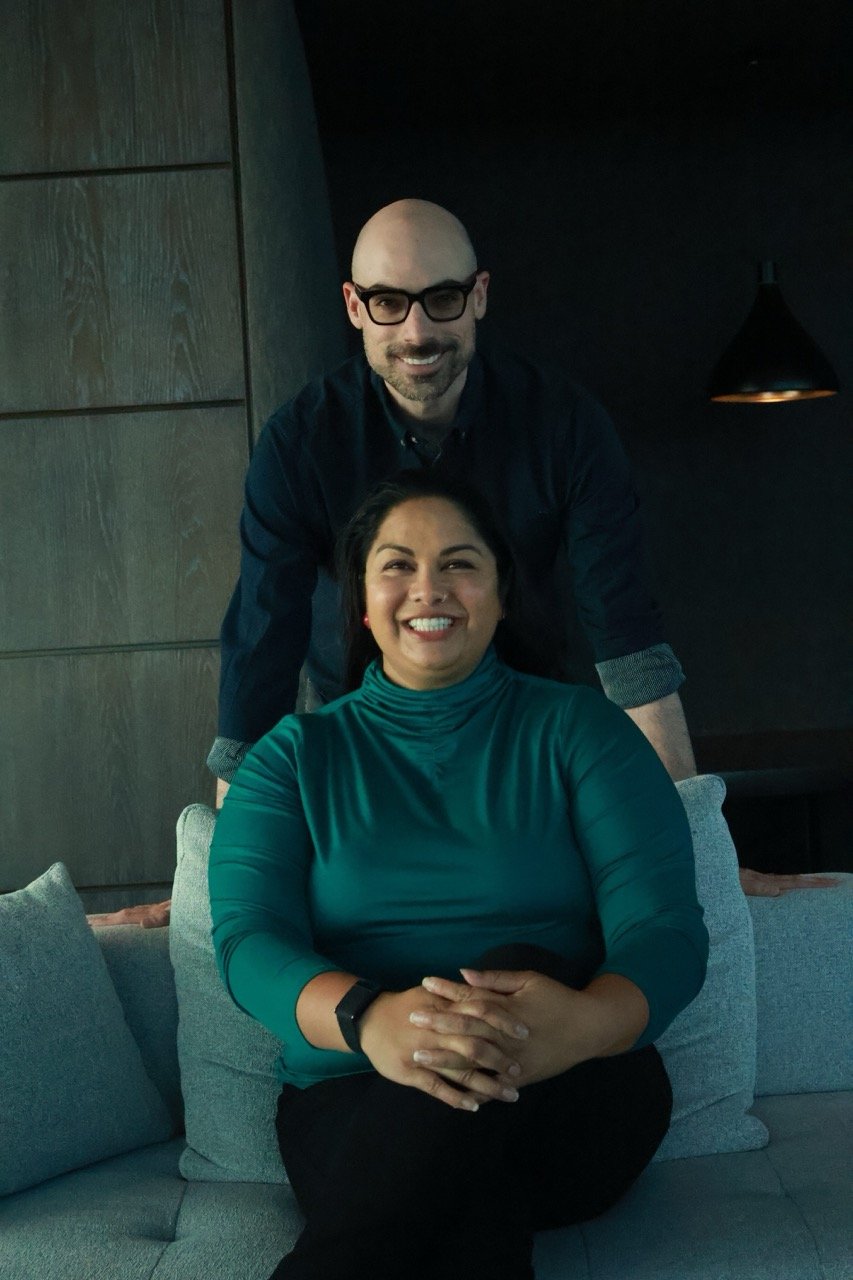 Boldly Go Beyond co-founders Zainab and Marc; Zainab is seated in a green turtleneck with Marc standing behind her in a dark shirt and glasses.