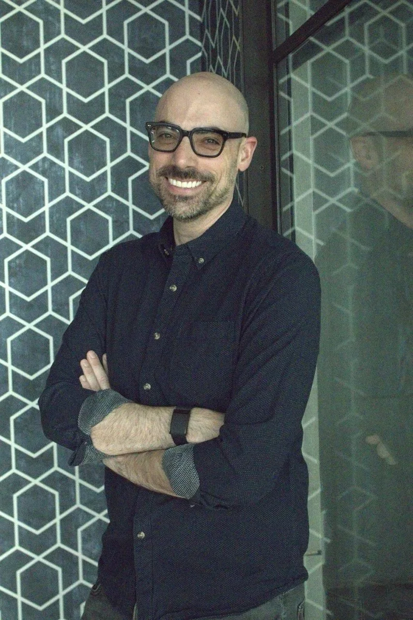 Co-founder Marc Chiu smiling and leaning against a wall with a geometric pattern. He is wearing glasses and a Whoop fitness tracker.
