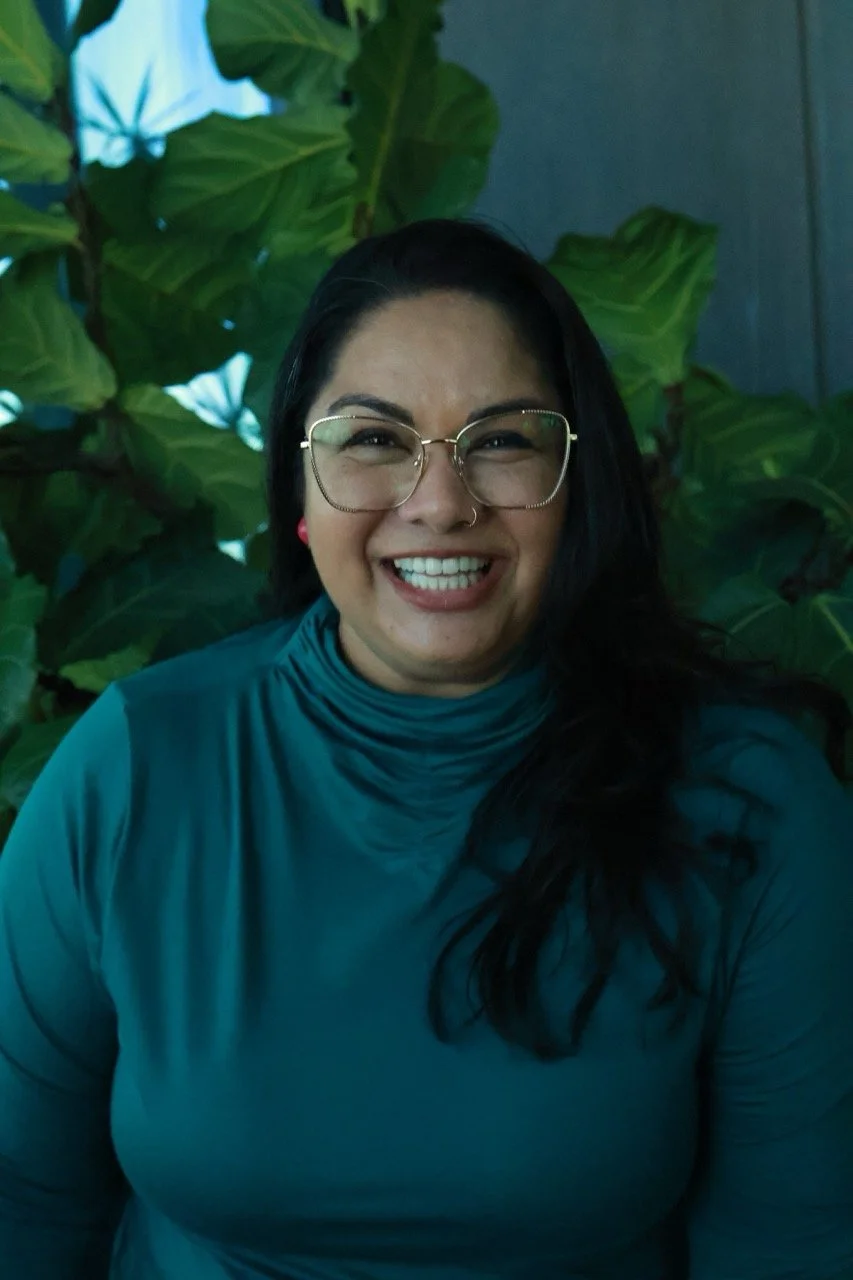 Co-founder Zainab Hafiz smiling and standing in front of  large plant wearing glasses and a green turtleneck and pink earrings.