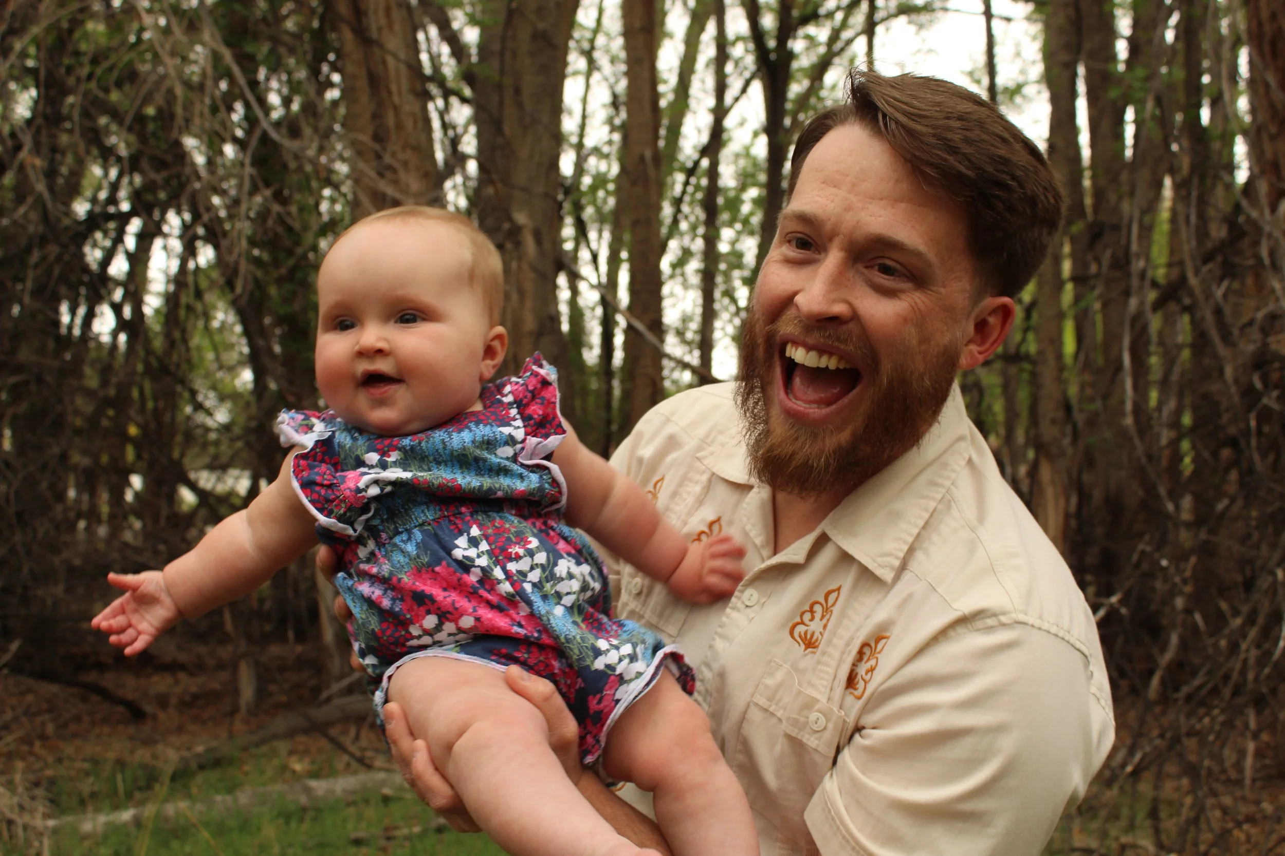 A man holding a smiling baby wearing a floral dress in a wooded area.
