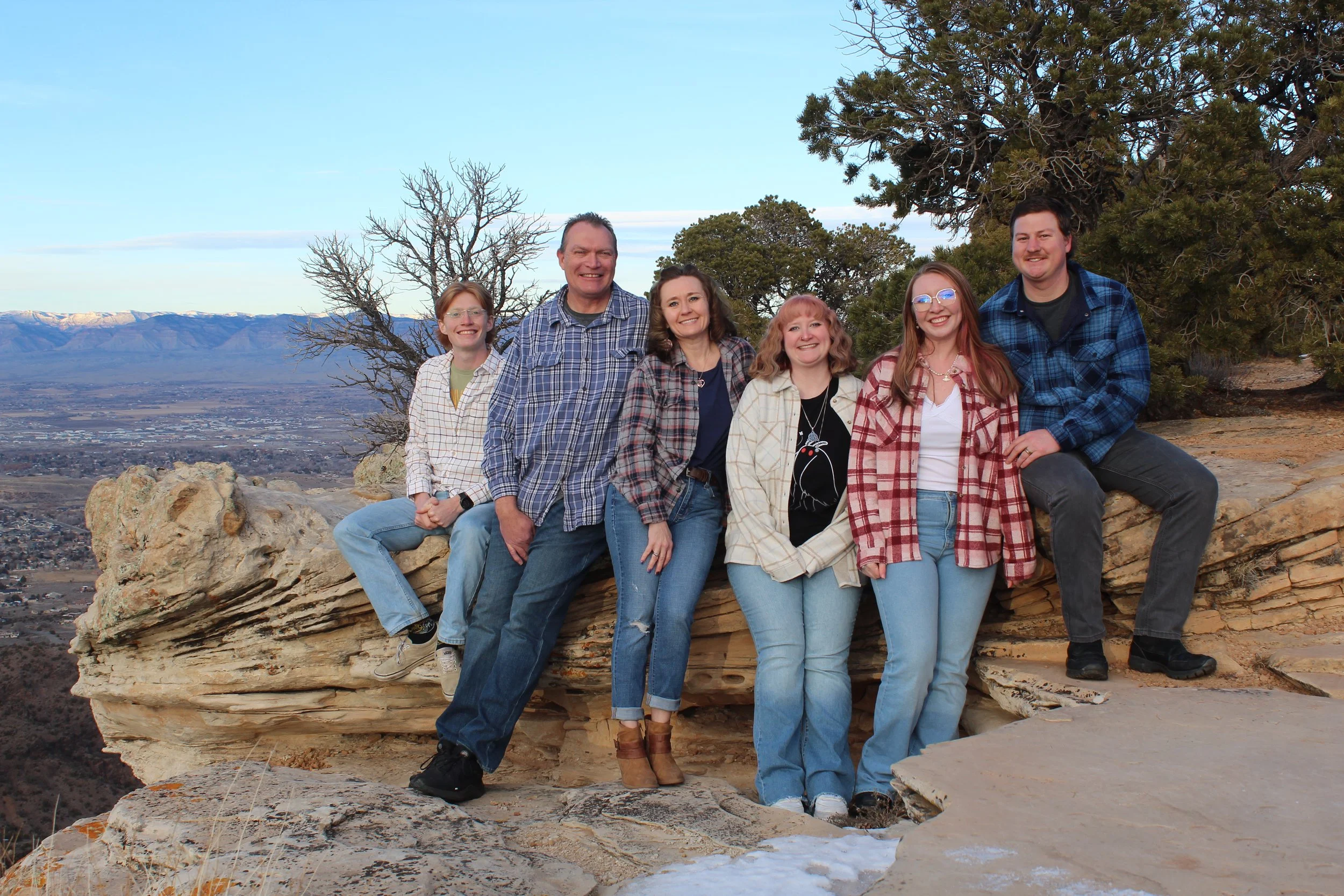 Group of six people sitting on a rocky ledge with a scenic mountainous landscape in the background, wearing casual plaid shirts and jeans.