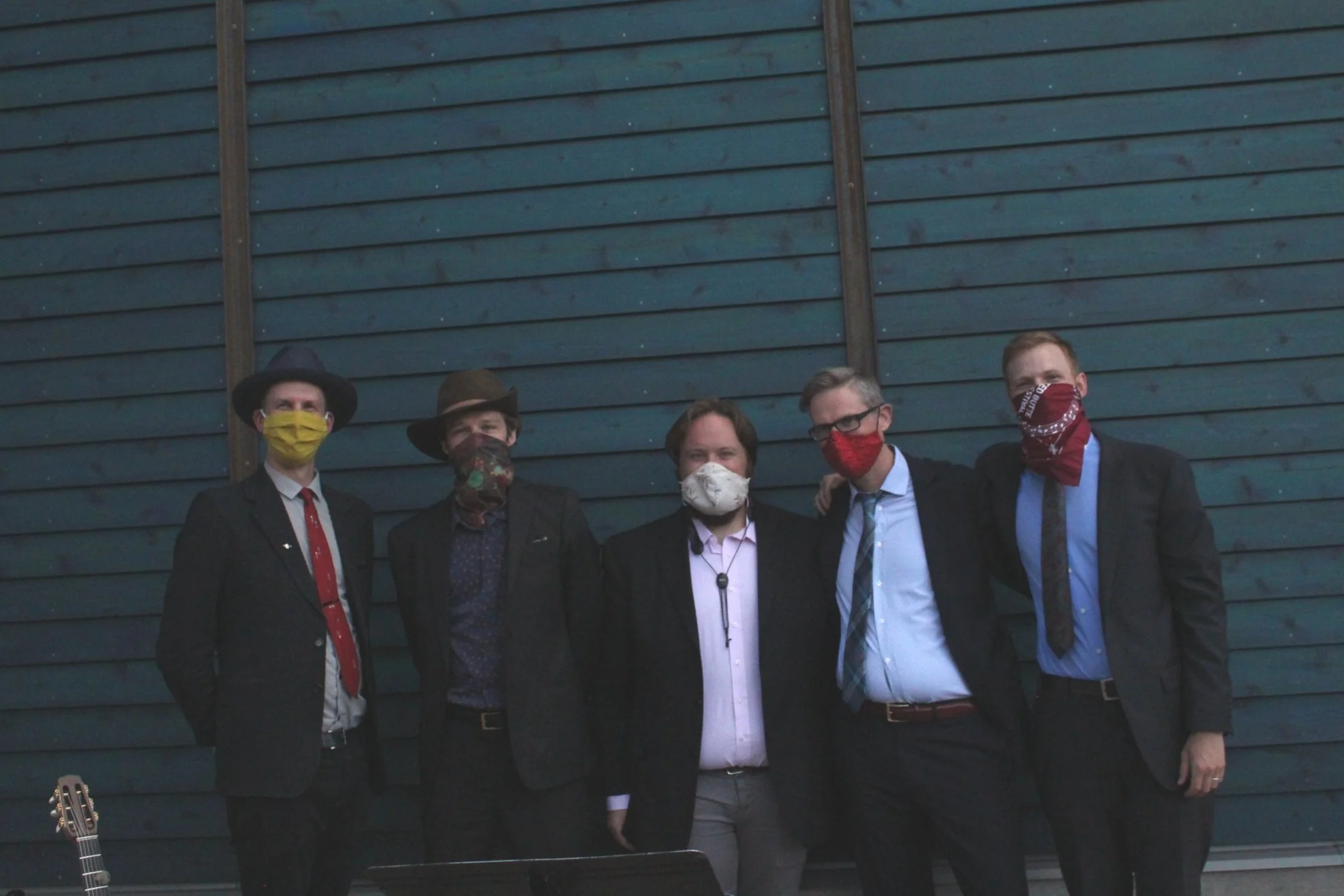 Five men wearing suits and face masks, posing in front of a blue wall. Some are also wearing hats.