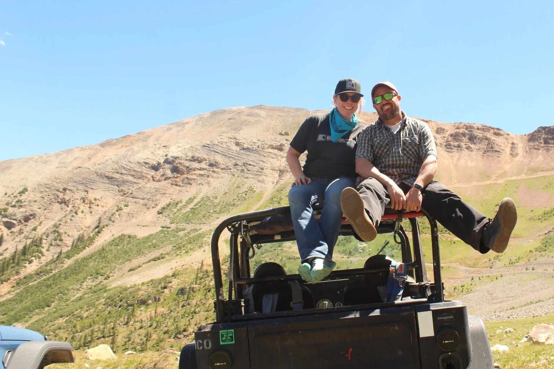 Two people sitting on a vehicle in a mountain landscape under a clear blue sky.