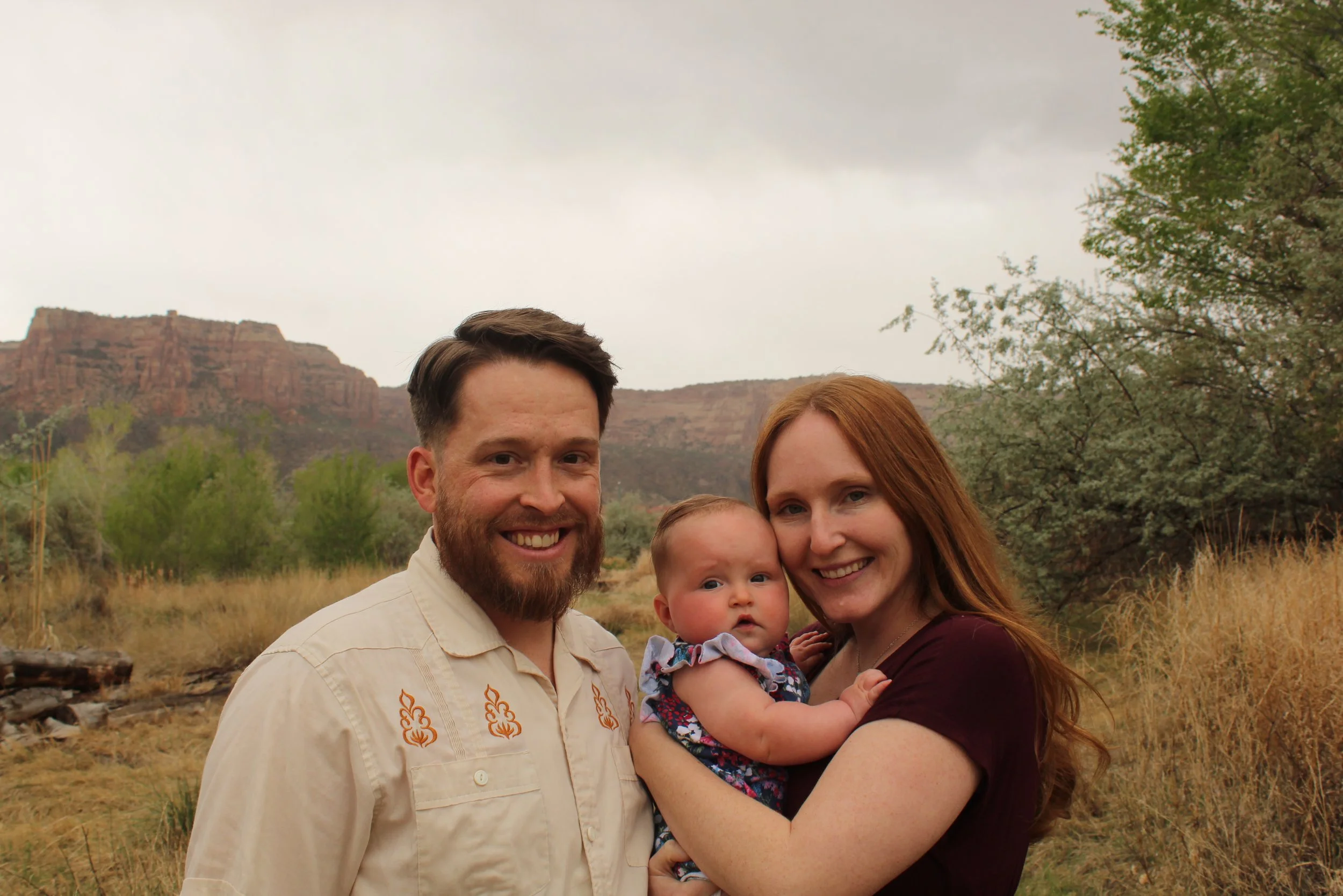 A couple holding a baby standing outdoors with a mountainous landscape background.