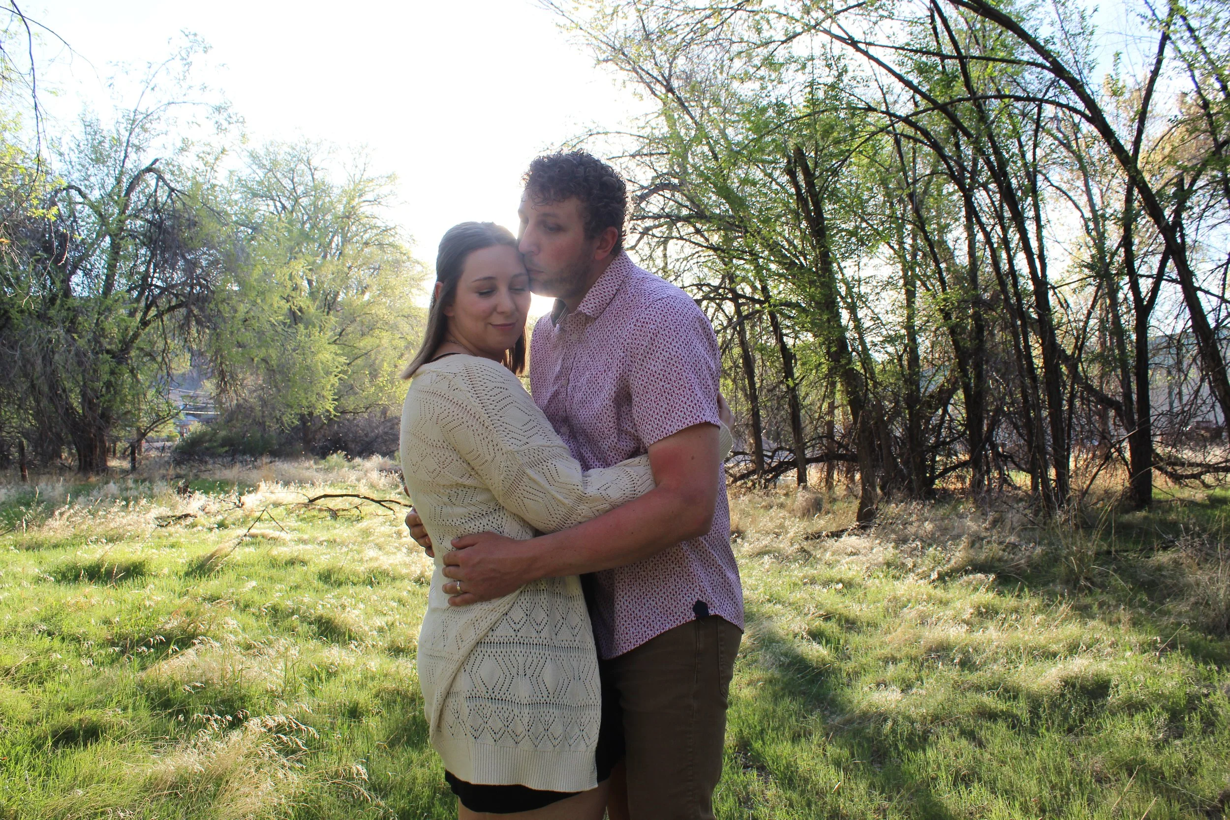 A couple embraces in a grassy field with trees in the background during daylight.