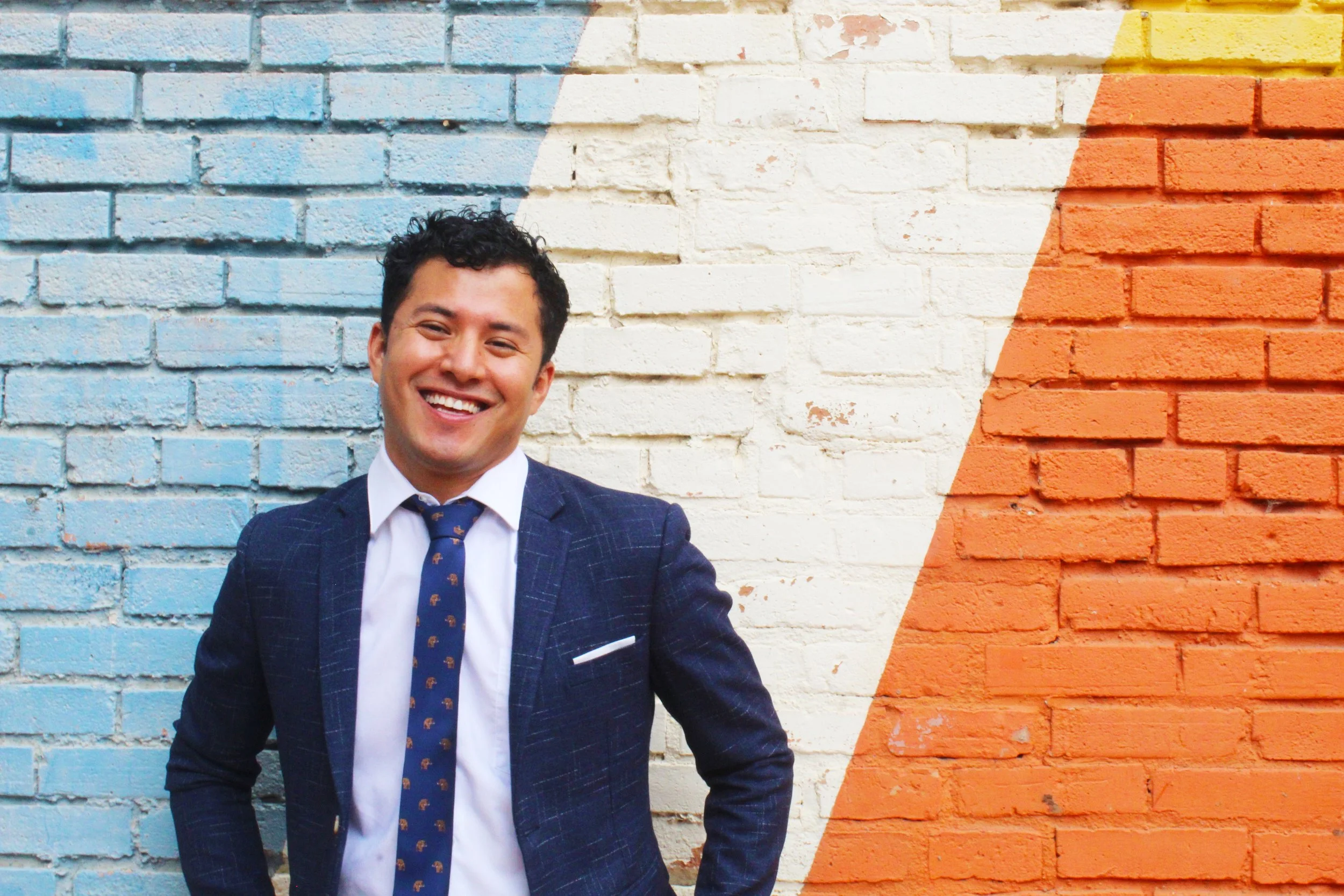 Man in a suit smiling in front of a colorful brick wall.