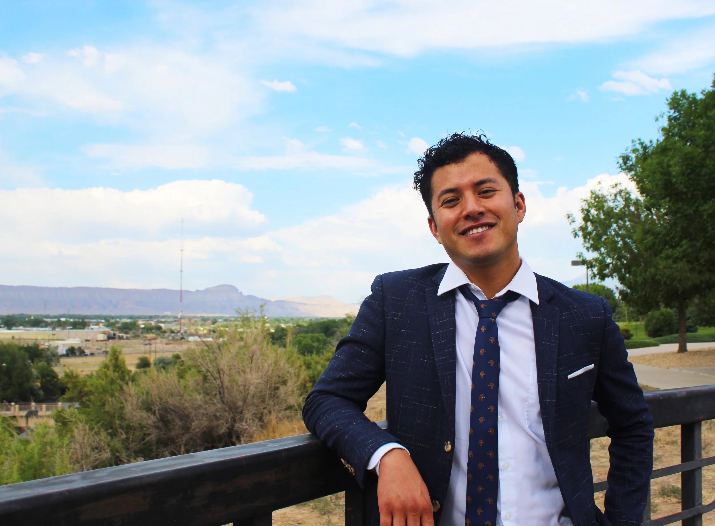 Smiling man in a suit leaning on a railing with a scenic landscape background.