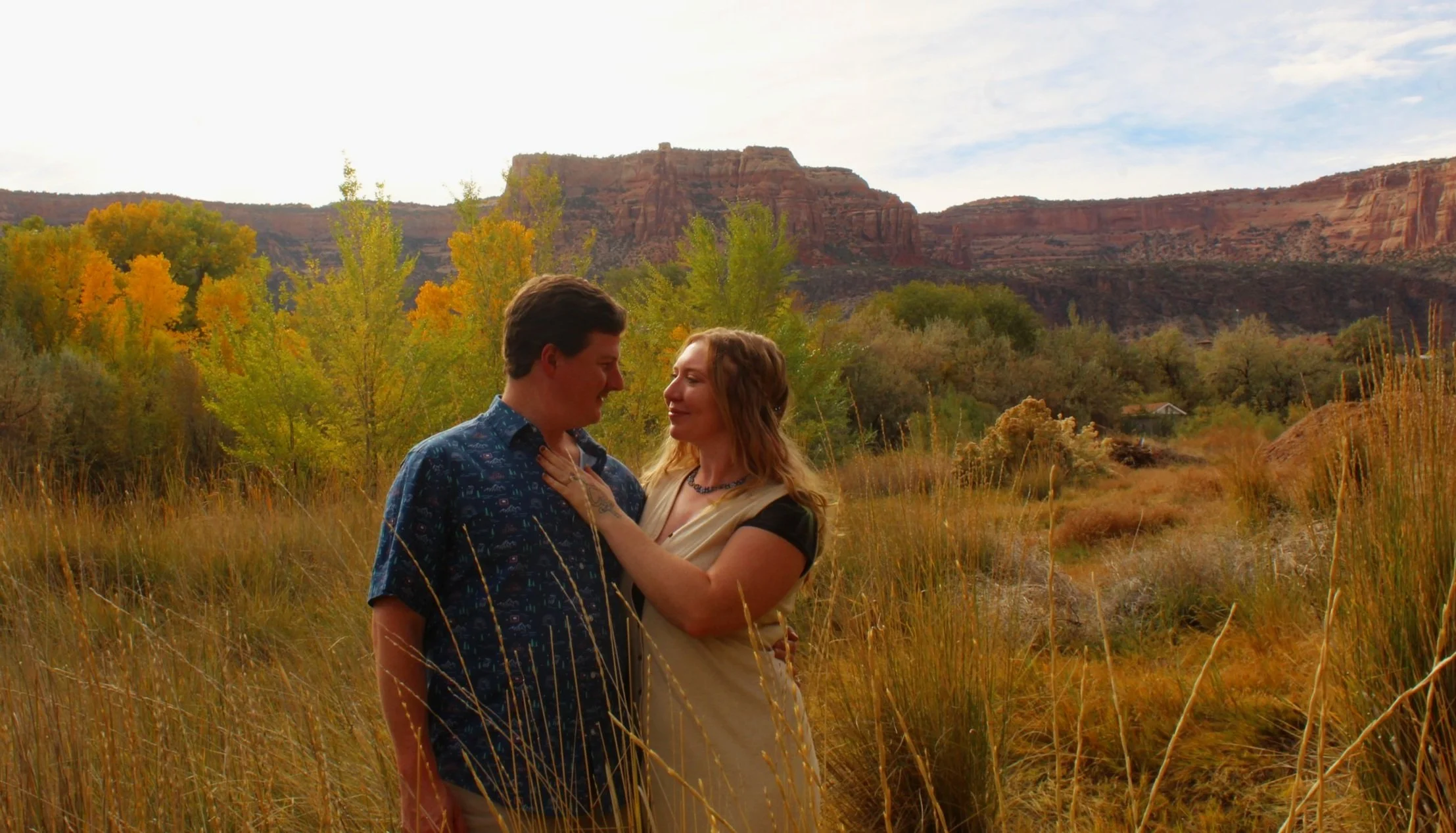 A couple standing in a field with autumn foliage and a rocky cliff in the background.