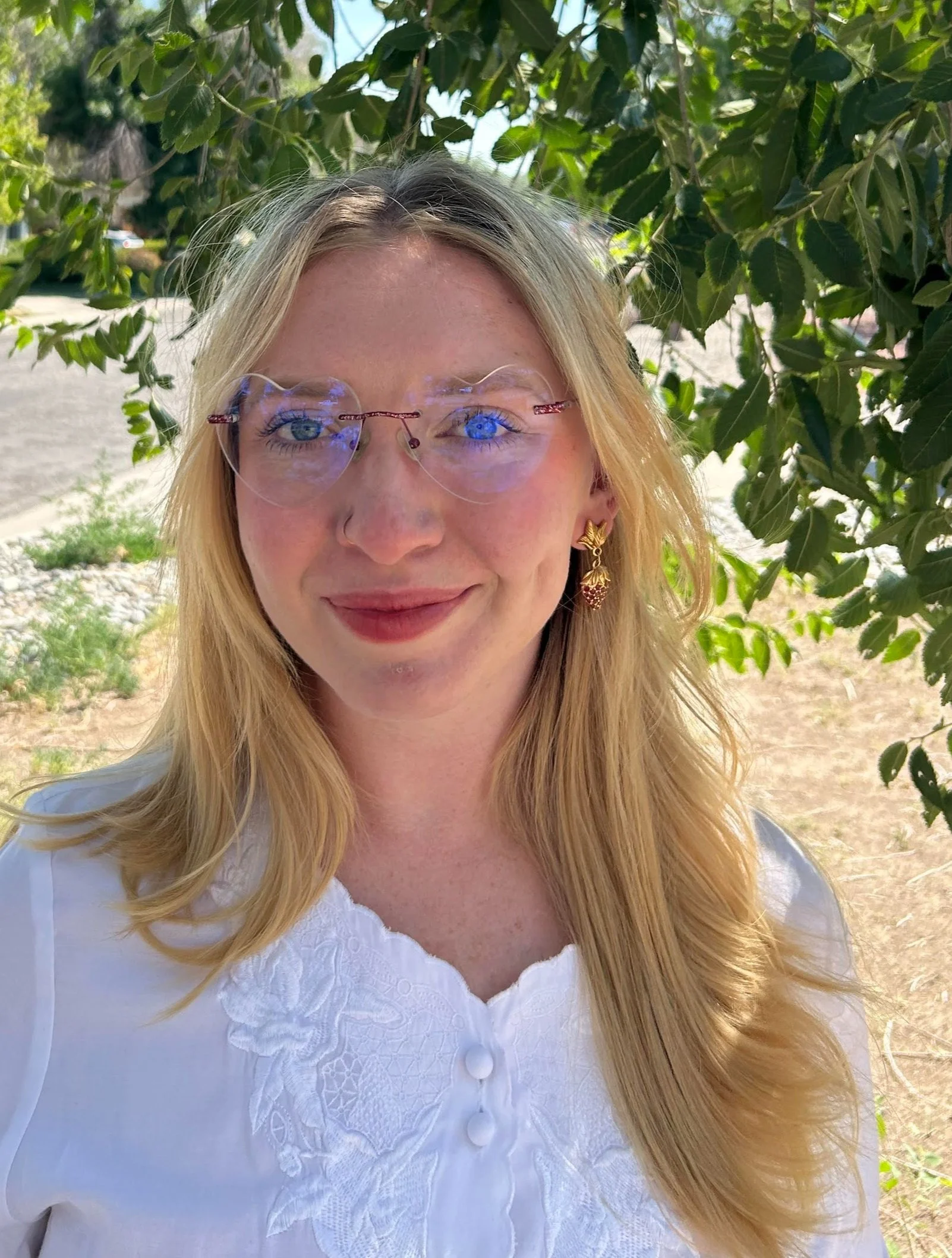 Woman with blonde hair, glasses, and a white blouse, standing outdoors with green foliage in the background.