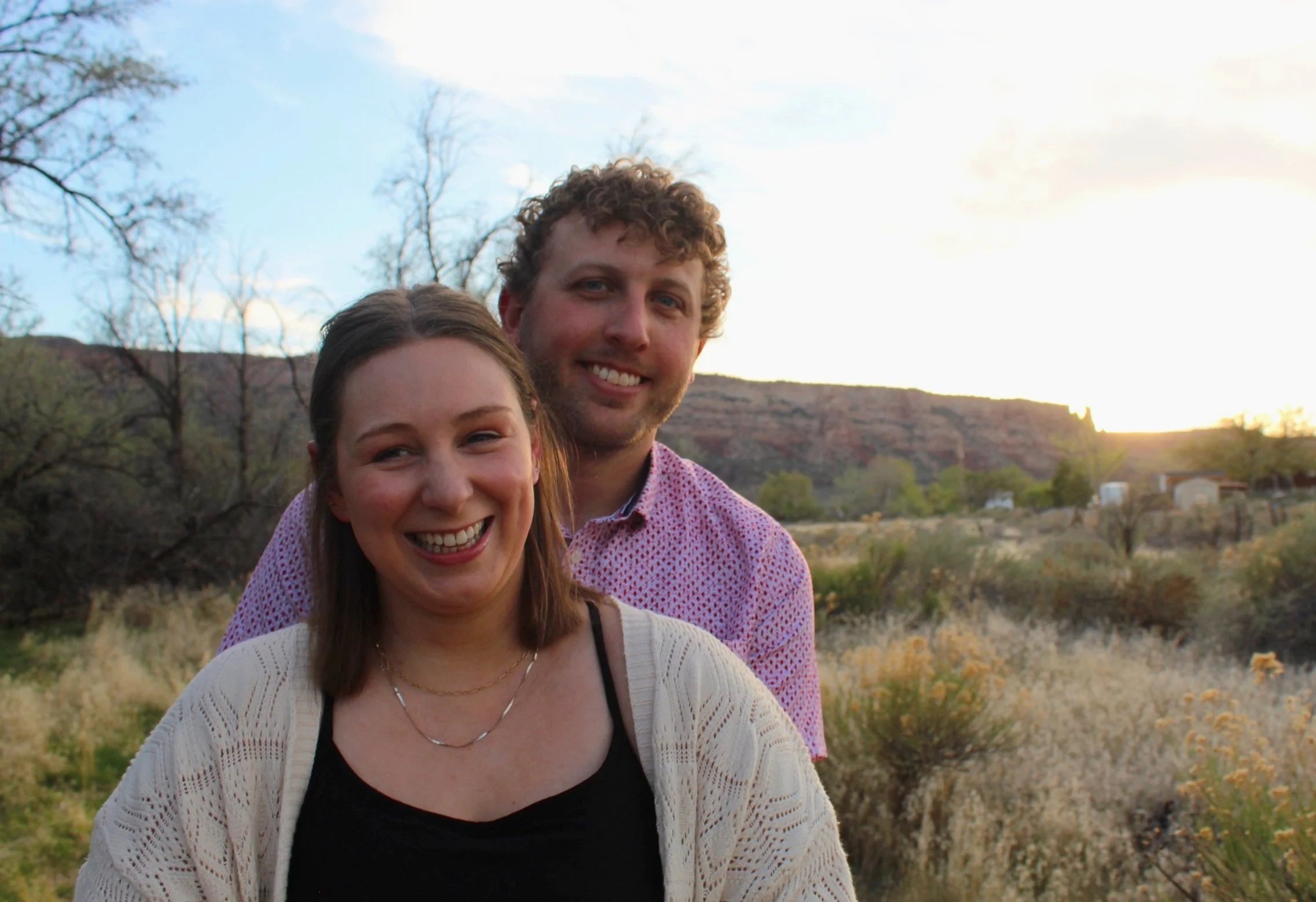 A man and woman smiling in an outdoor setting with dry vegetation and a sunset in the background.