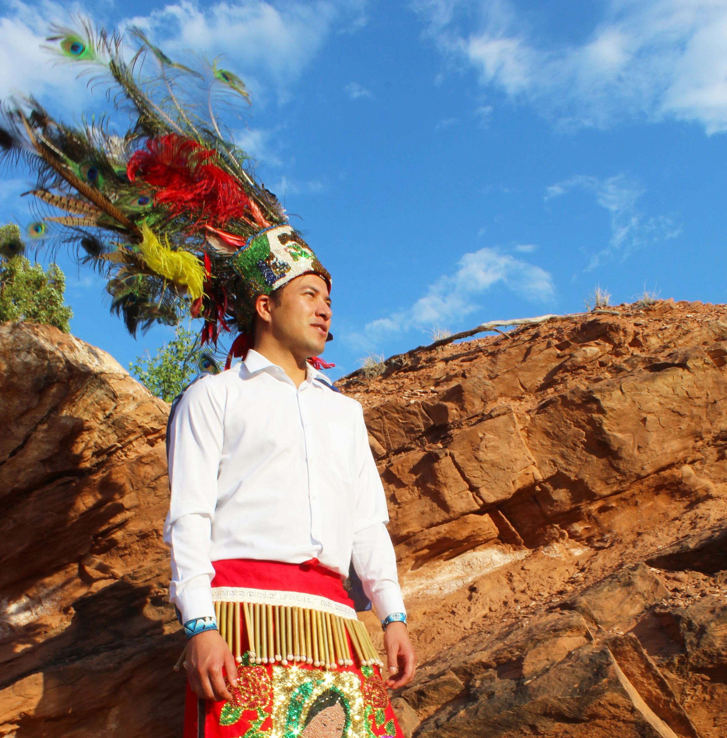 Person in traditional costume with a feathered headdress standing against a rock formation and blue sky.