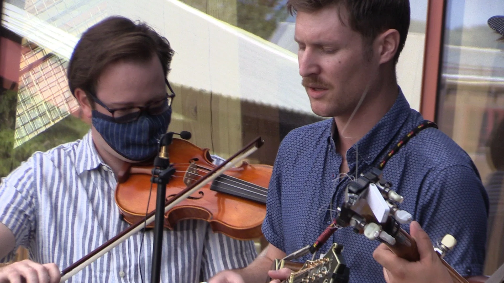 Two musicians performing with a violin and a string instrument, one wearing a face mask, outdoor setting.