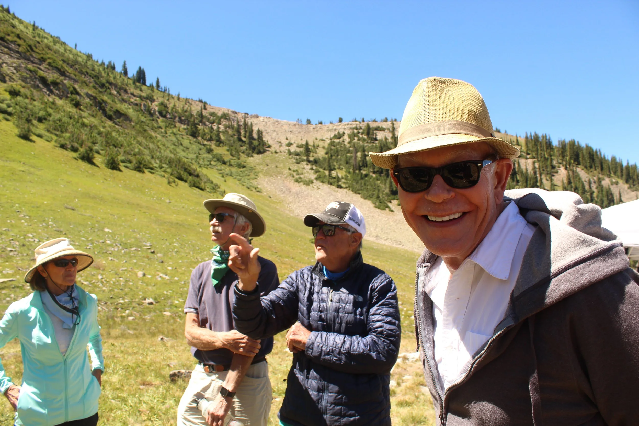 Group of four people outdoors in a grassy hilly area, wearing hats and sunglasses on a sunny day.