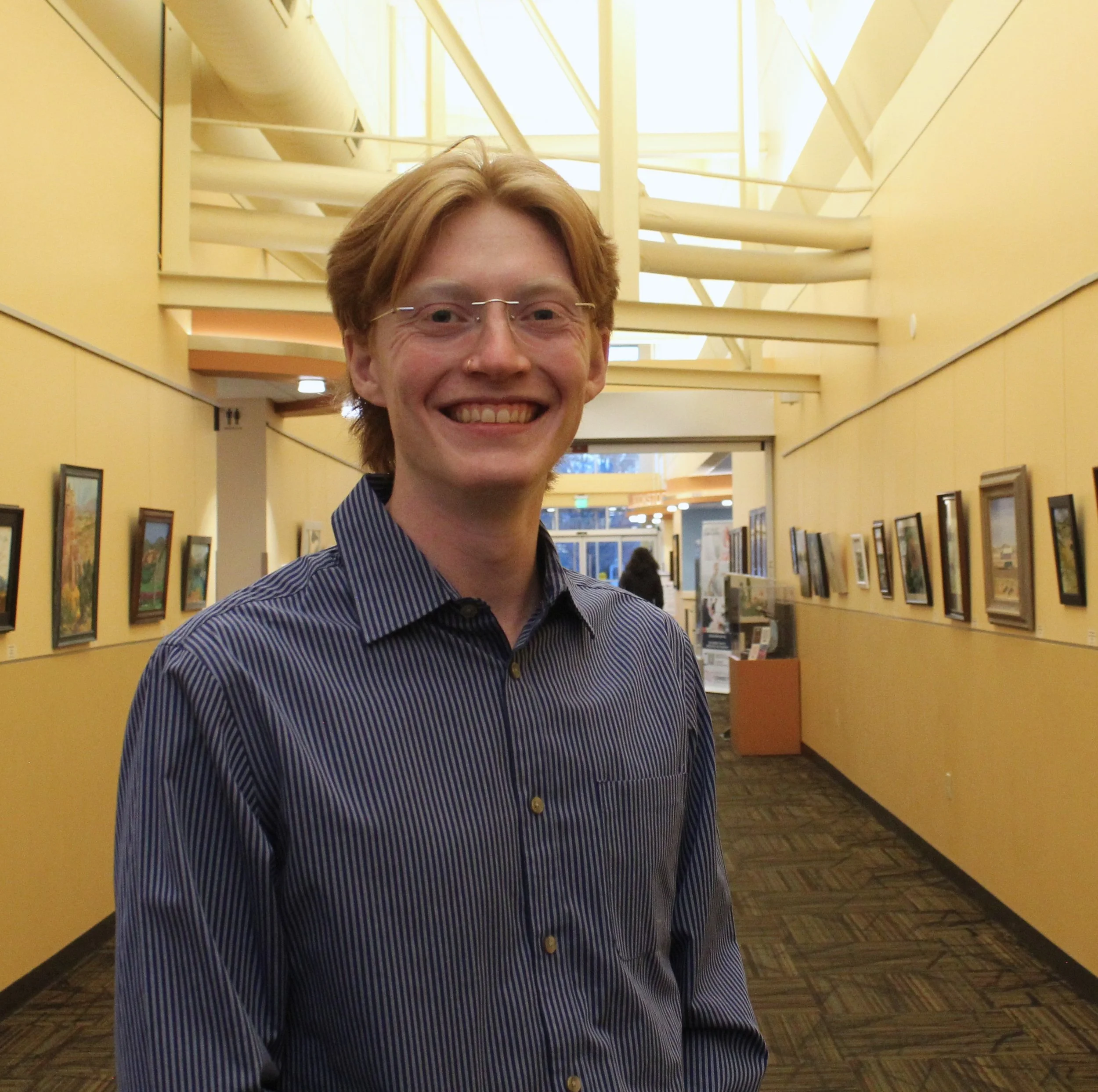 Smiling person in a hallway with paintings on the walls, wearing a striped shirt.
