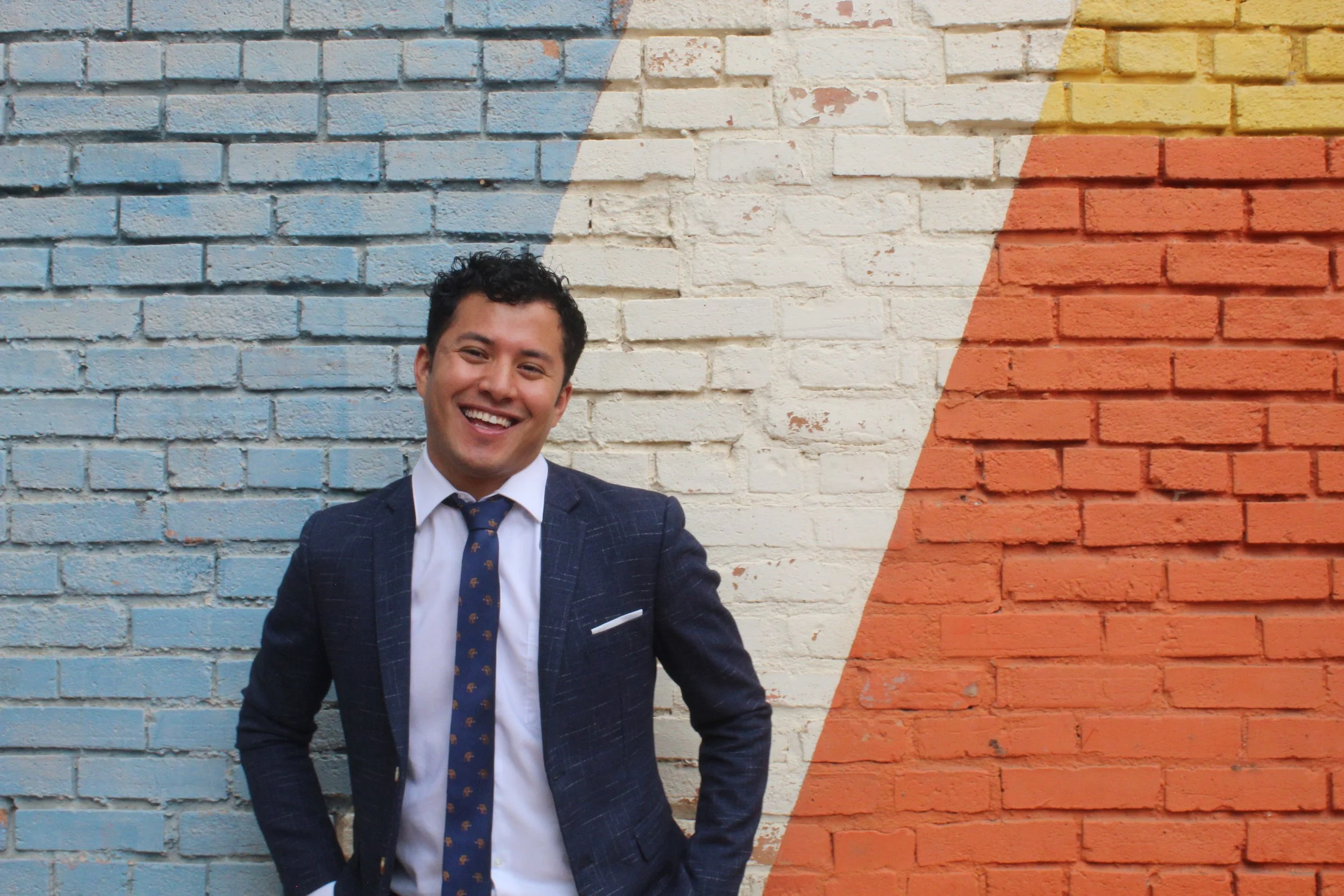 Man in a suit standing in front of a colorful brick wall with blue, white, orange, and yellow sections.