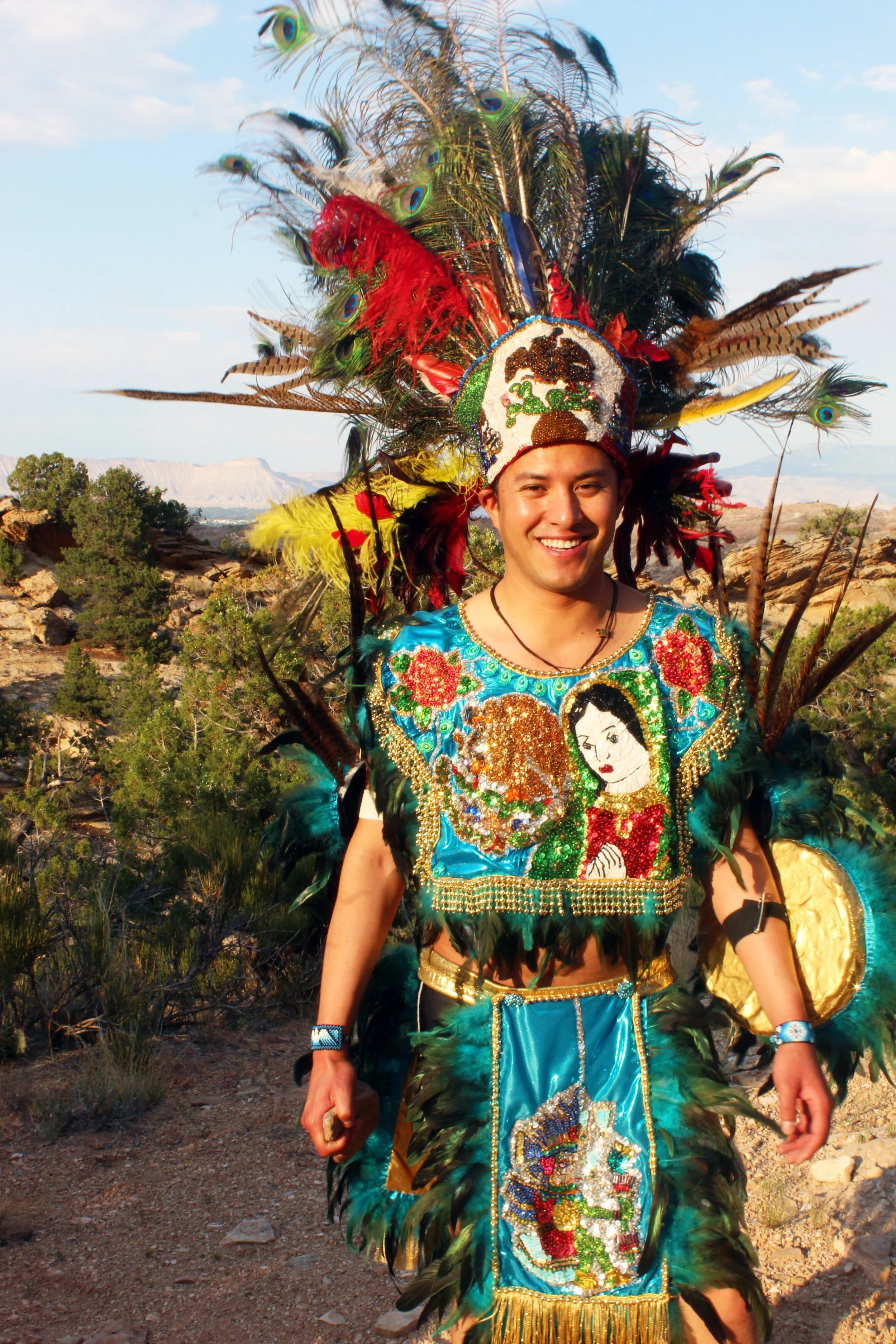 Person wearing colorful traditional costume with feathers and intricate beadwork, standing outdoors in a natural landscape.