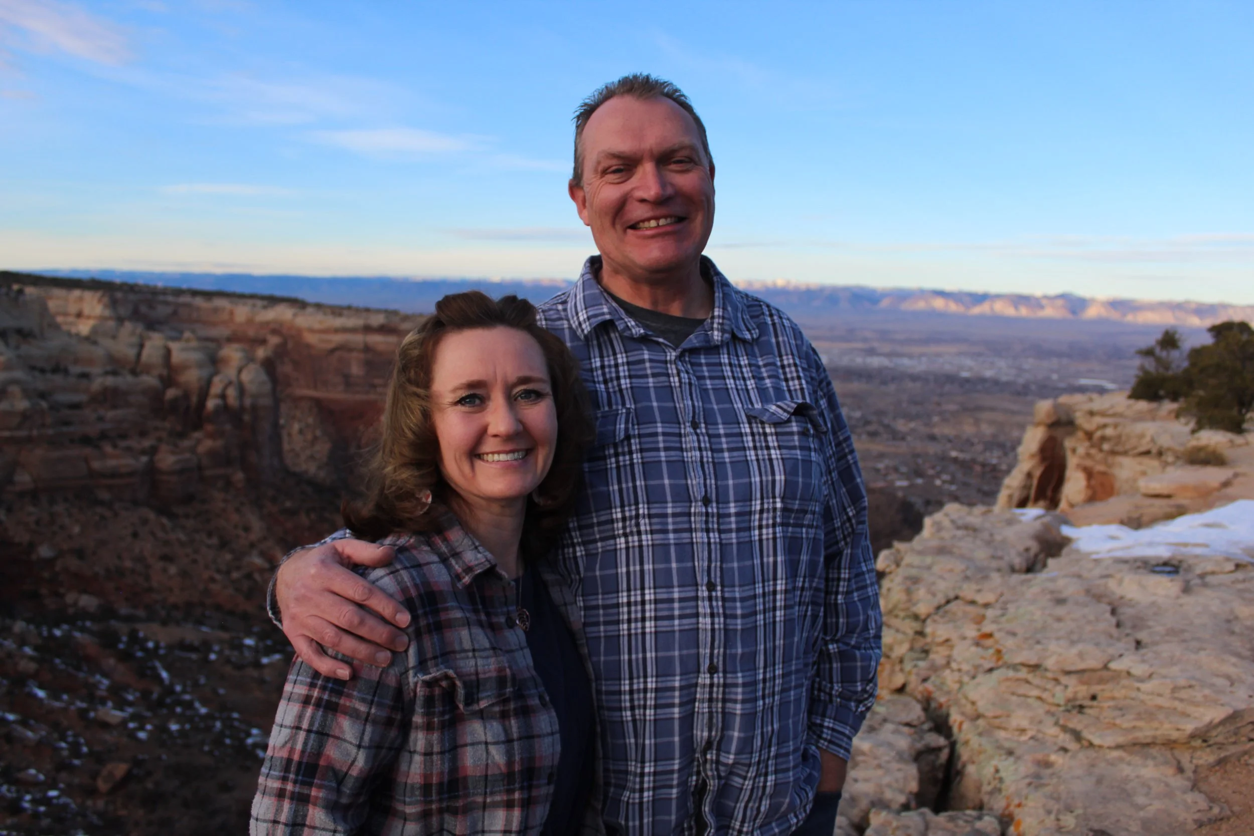 A man and woman in plaid shirts stand smiling on a rocky cliff with a vast canyon landscape in the background under a blue sky.