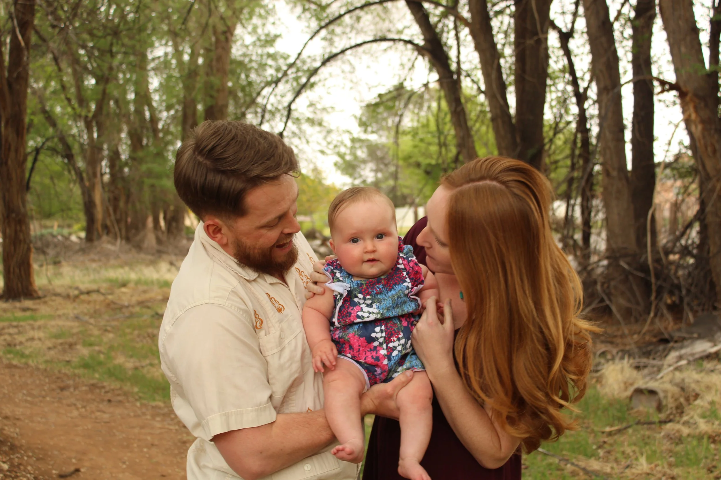 A couple holding a baby in a wooded area, with greenery and trees in the background. The man is wearing a light-colored shirt, and the woman has long red hair.