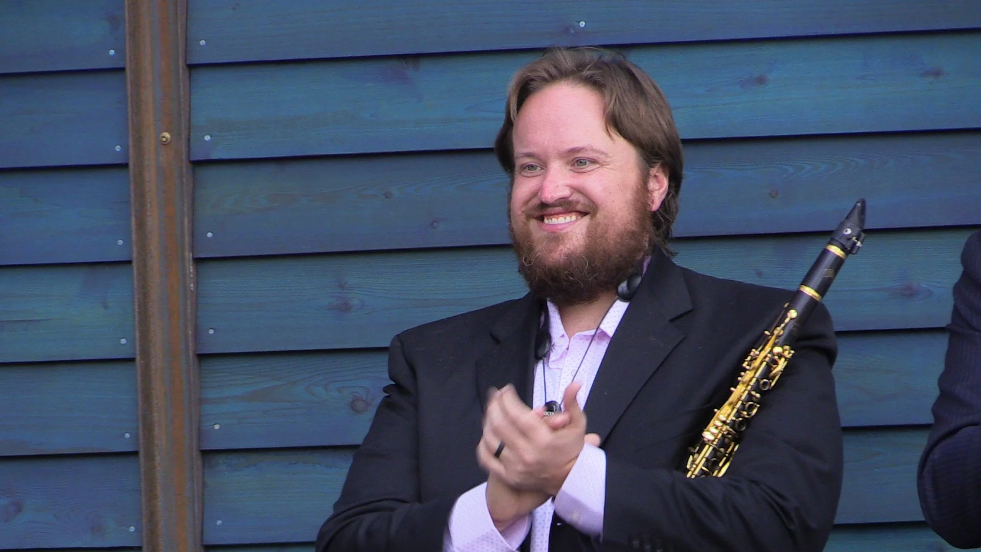 Man with instrument in hand, smiling and clapping, standing against blue wooden wall.