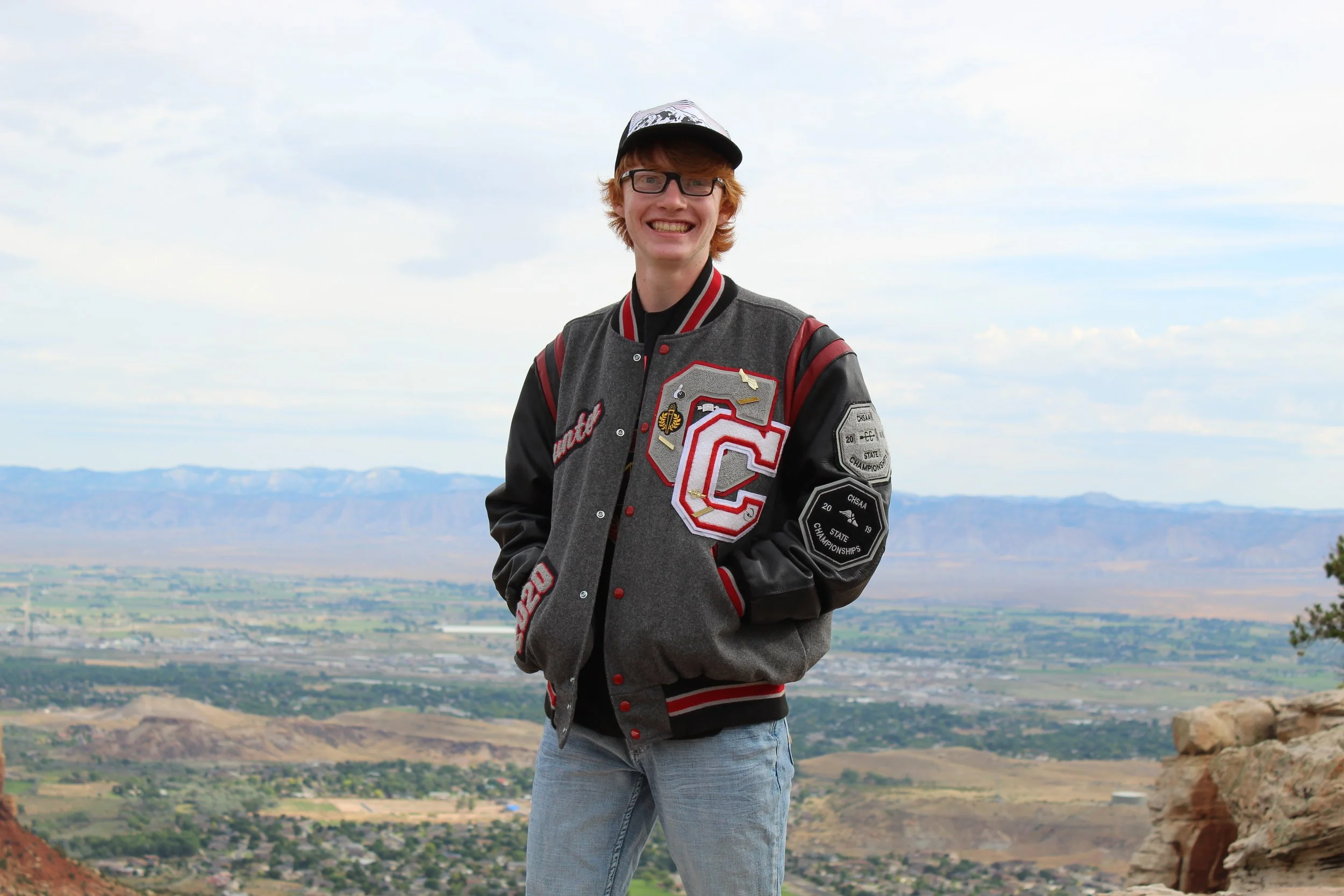 Smiling person wearing a letterman jacket and cap, standing outdoors with a scenic mountain and valley view in the background.