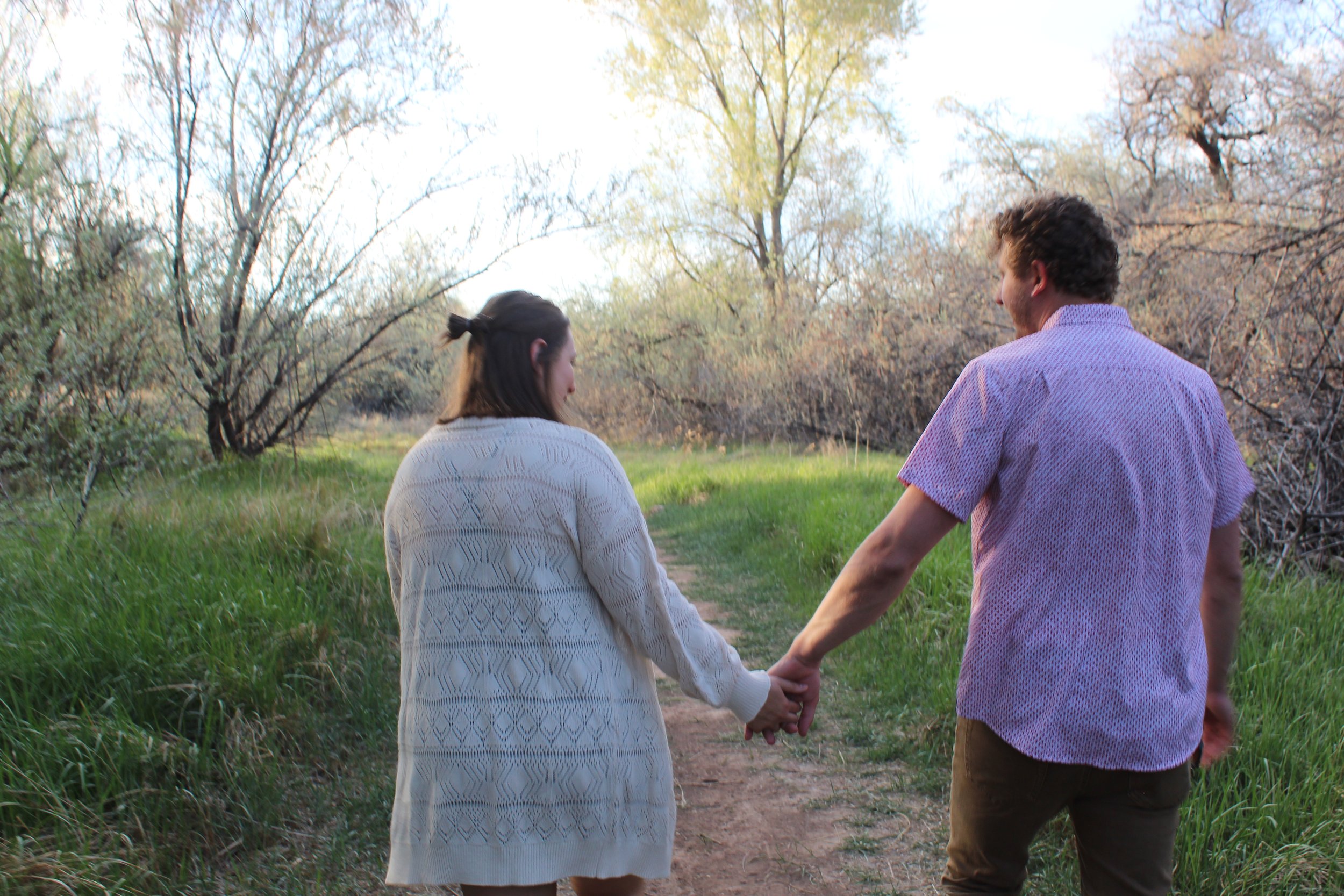 A couple holding hands while walking on a path in a leafy forest area.