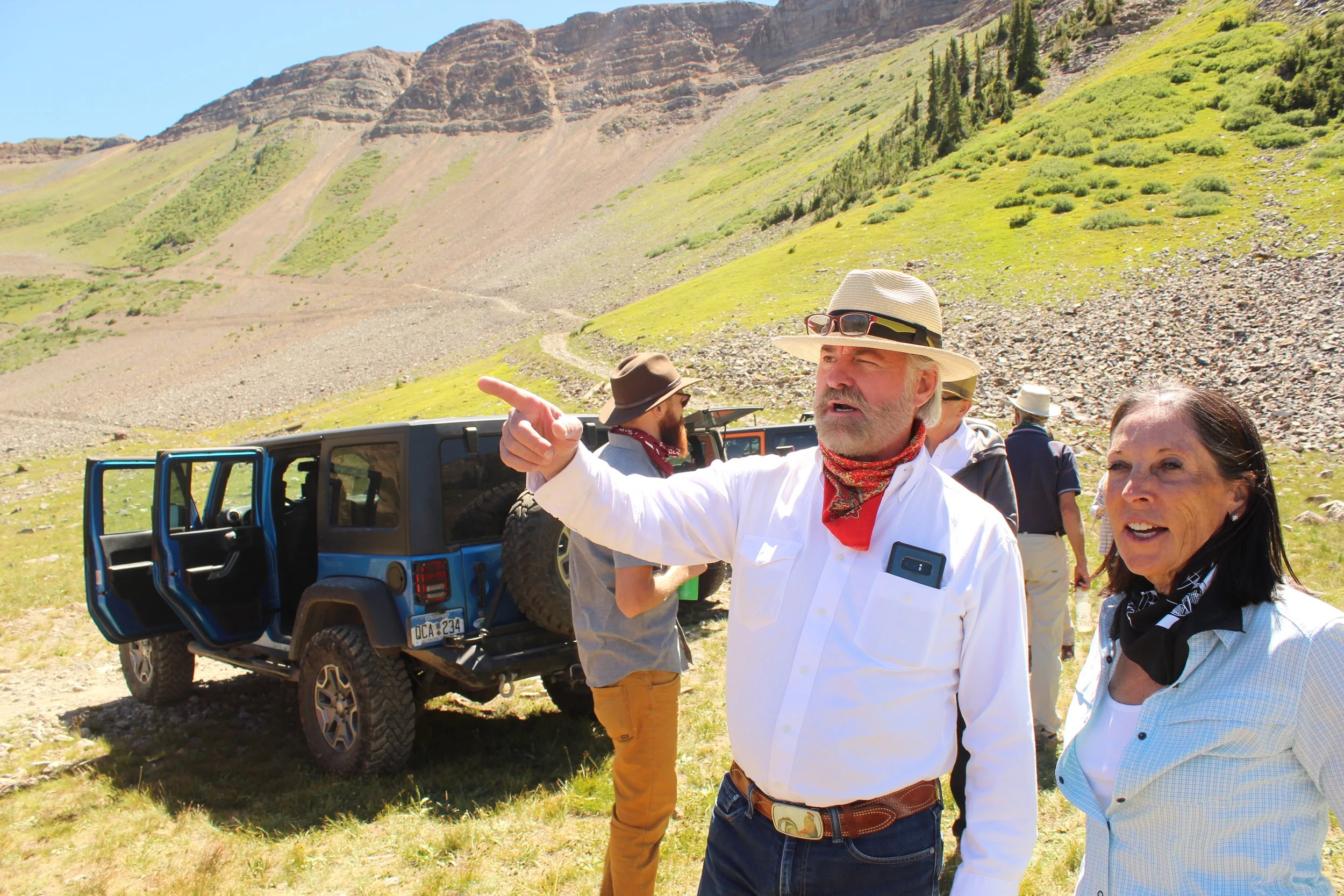 Group of people outdoors near a blue Jeep in a mountainous area, with a man in a hat pointing.