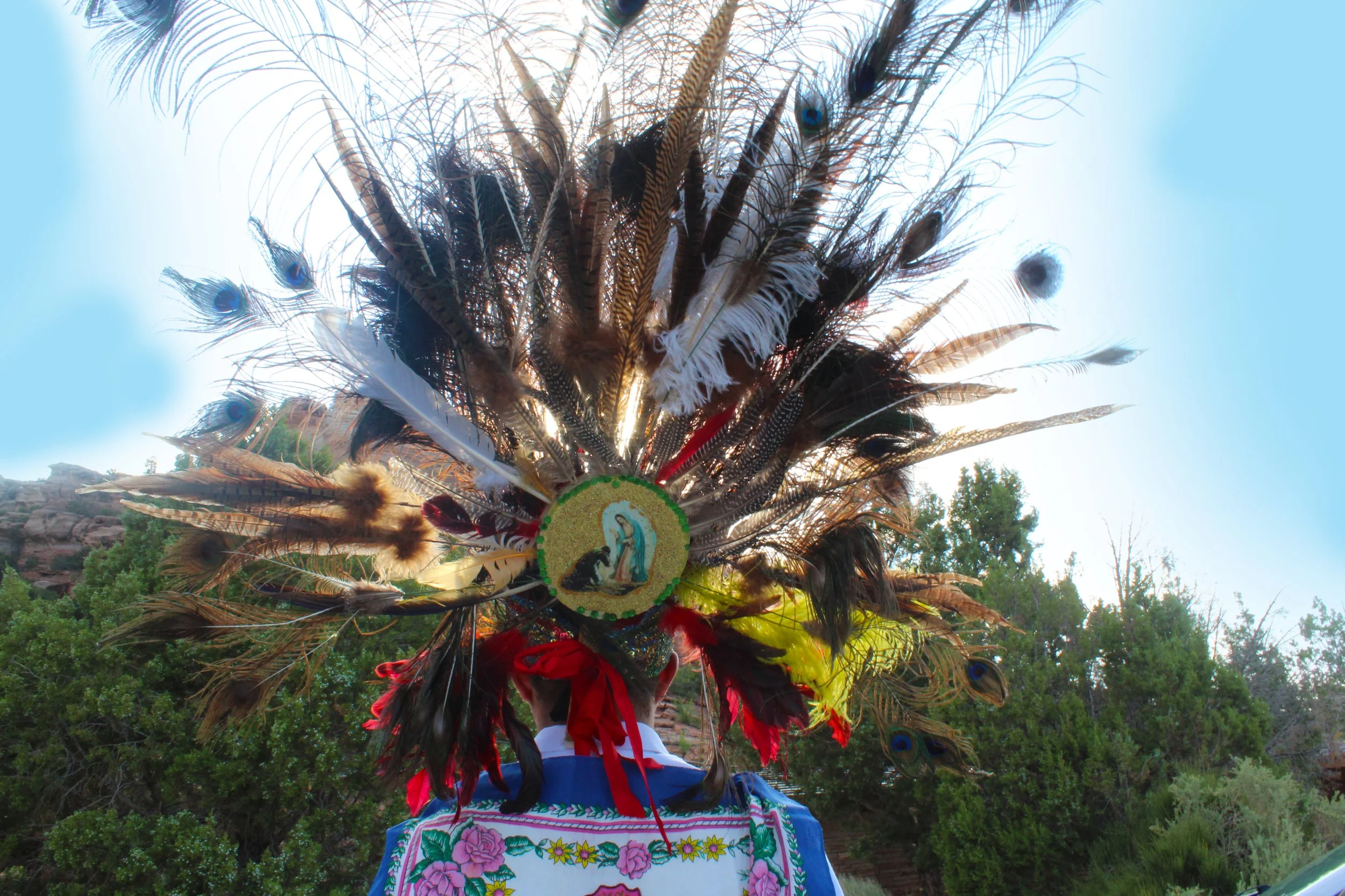 Person wearing a colorful feather headdress with vibrant patterns and embroidery, standing outdoors with trees and sky in the background.