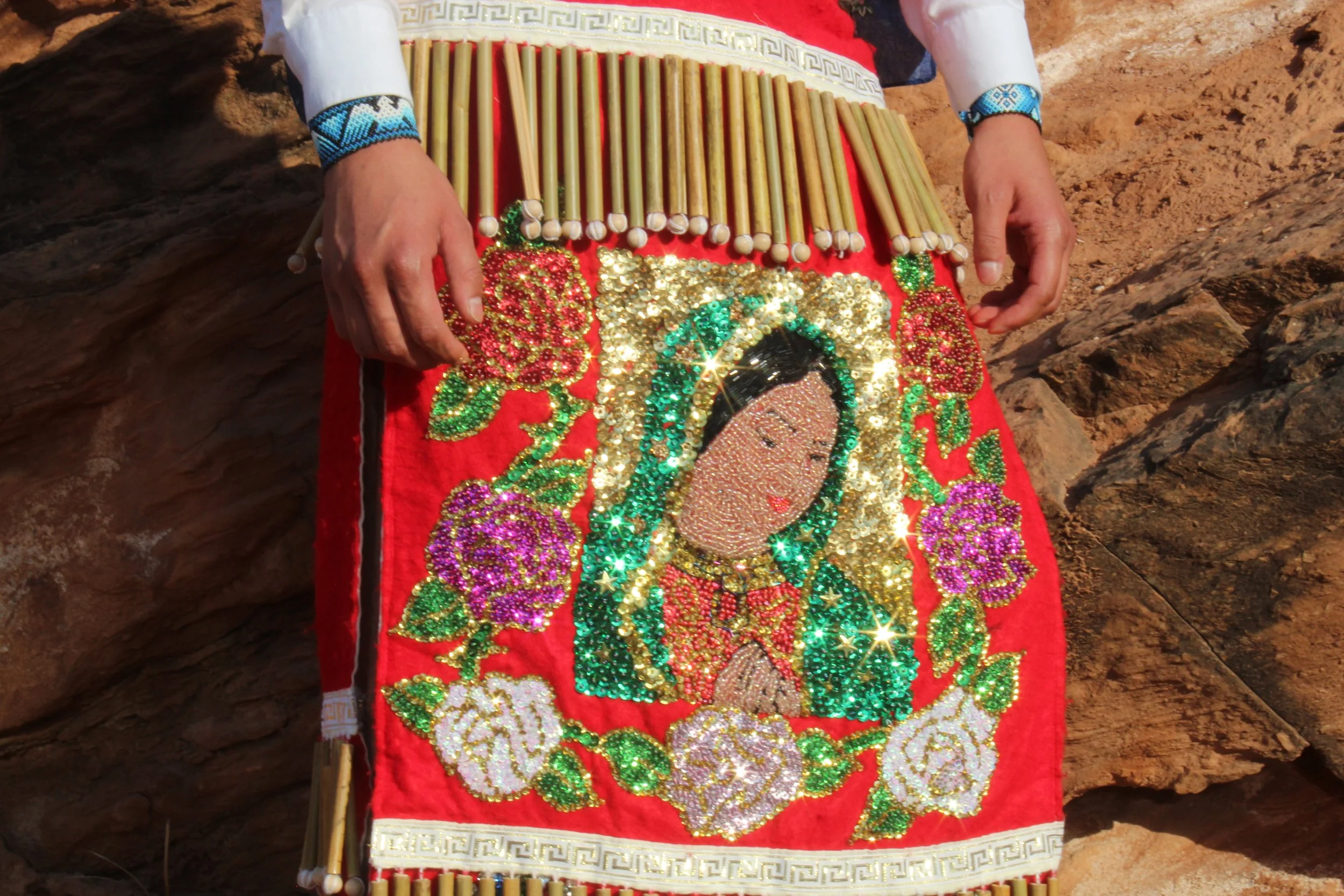 A decorative textile with a sequined image of a religious figure surrounded by colorful floral designs and bamboo sticks.