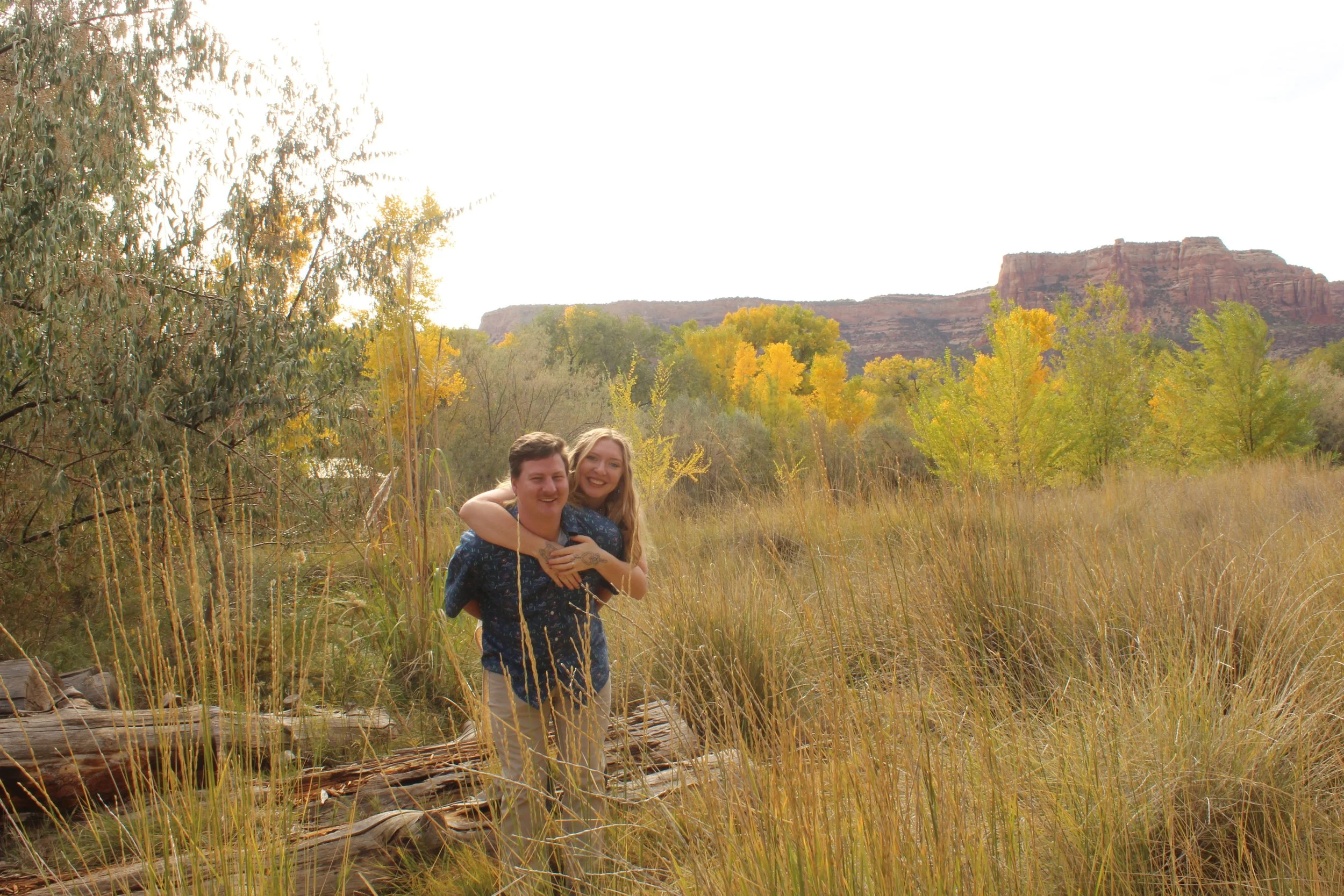 Couple playfully piggybacking in a grassy field with colorful autumn trees and red rock cliffs in the background.