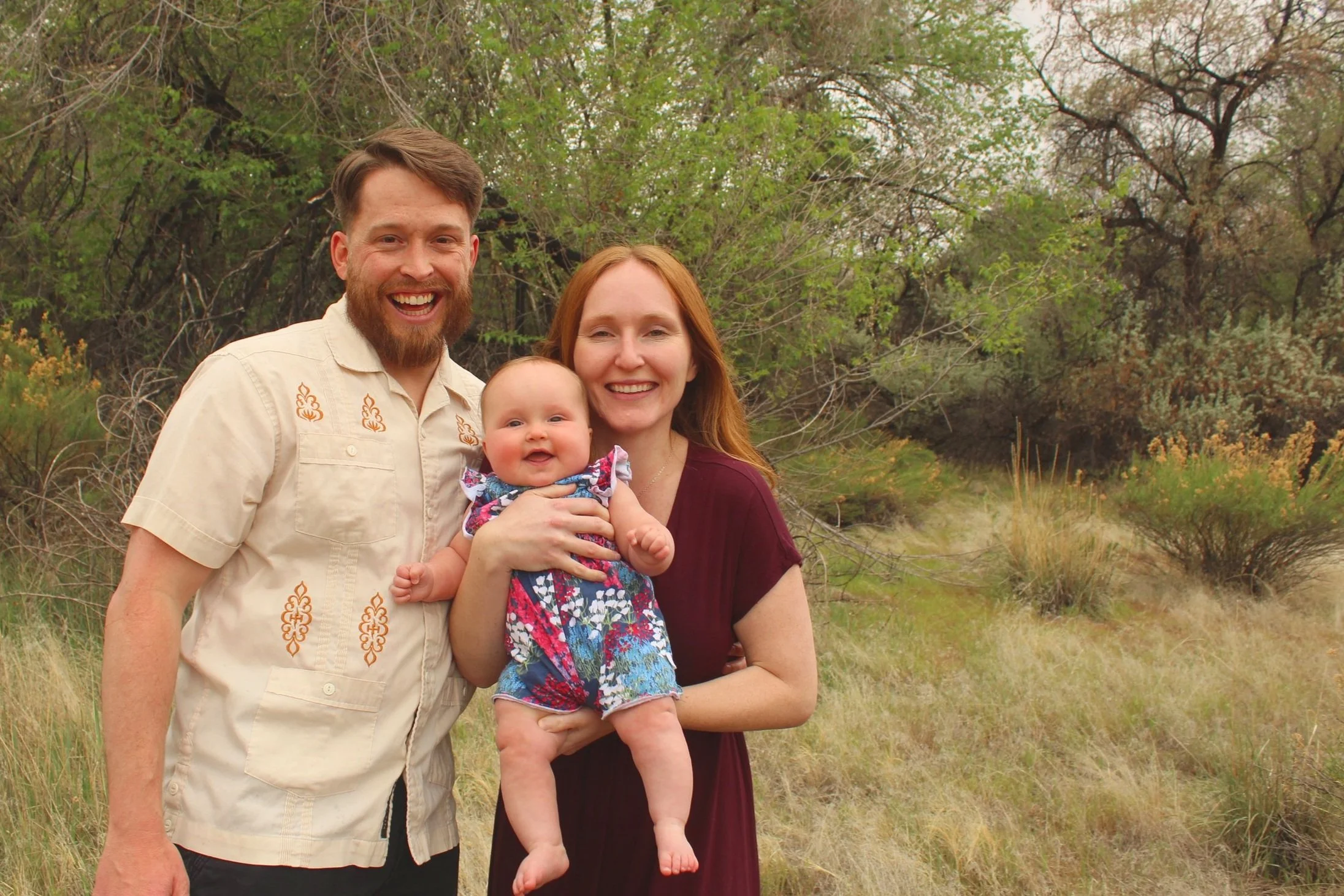 A smiling couple holding a baby in an outdoor setting with green trees and shrubs.
