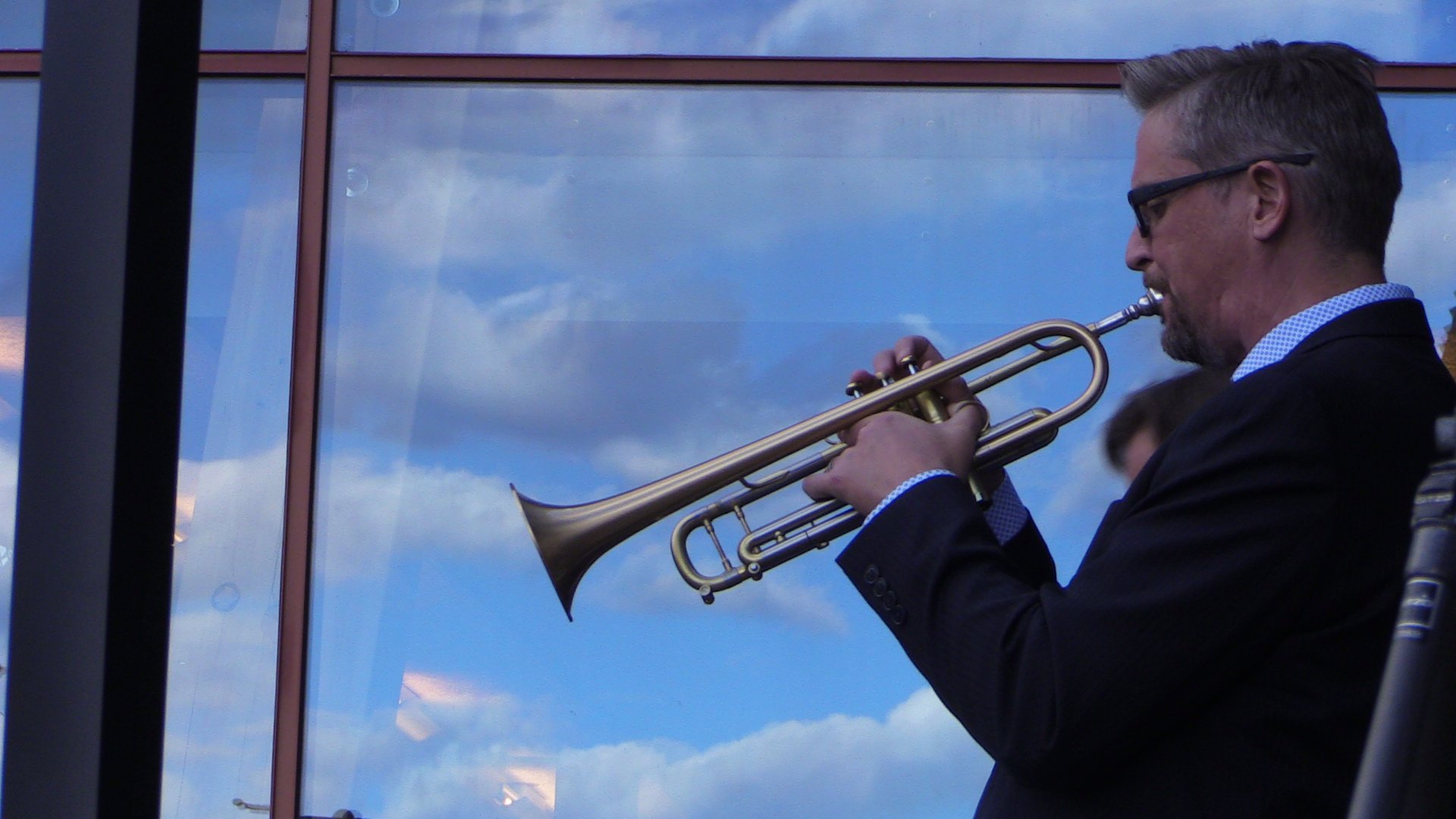 Man playing trumpet outside against blue sky and clouds reflection in glass.