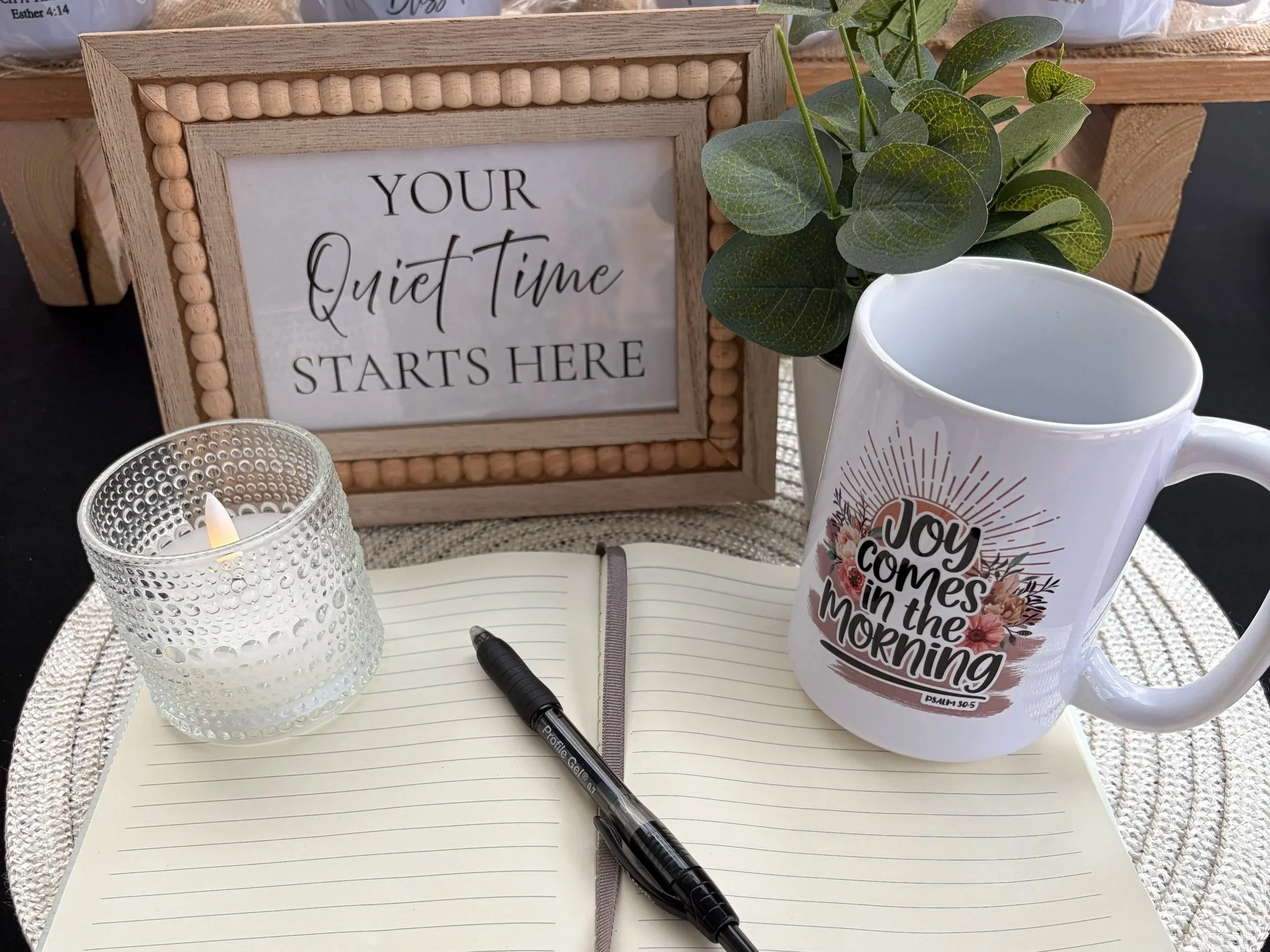 A framed sign with the message 'Your Quiet Time Starts Here' on a table. Nearby, there is a cup with a floral design and the phrase 'Joy Comes in the Morning,' a lit candle in a textured glass holder, an open notebook with a pen, and a green plant.