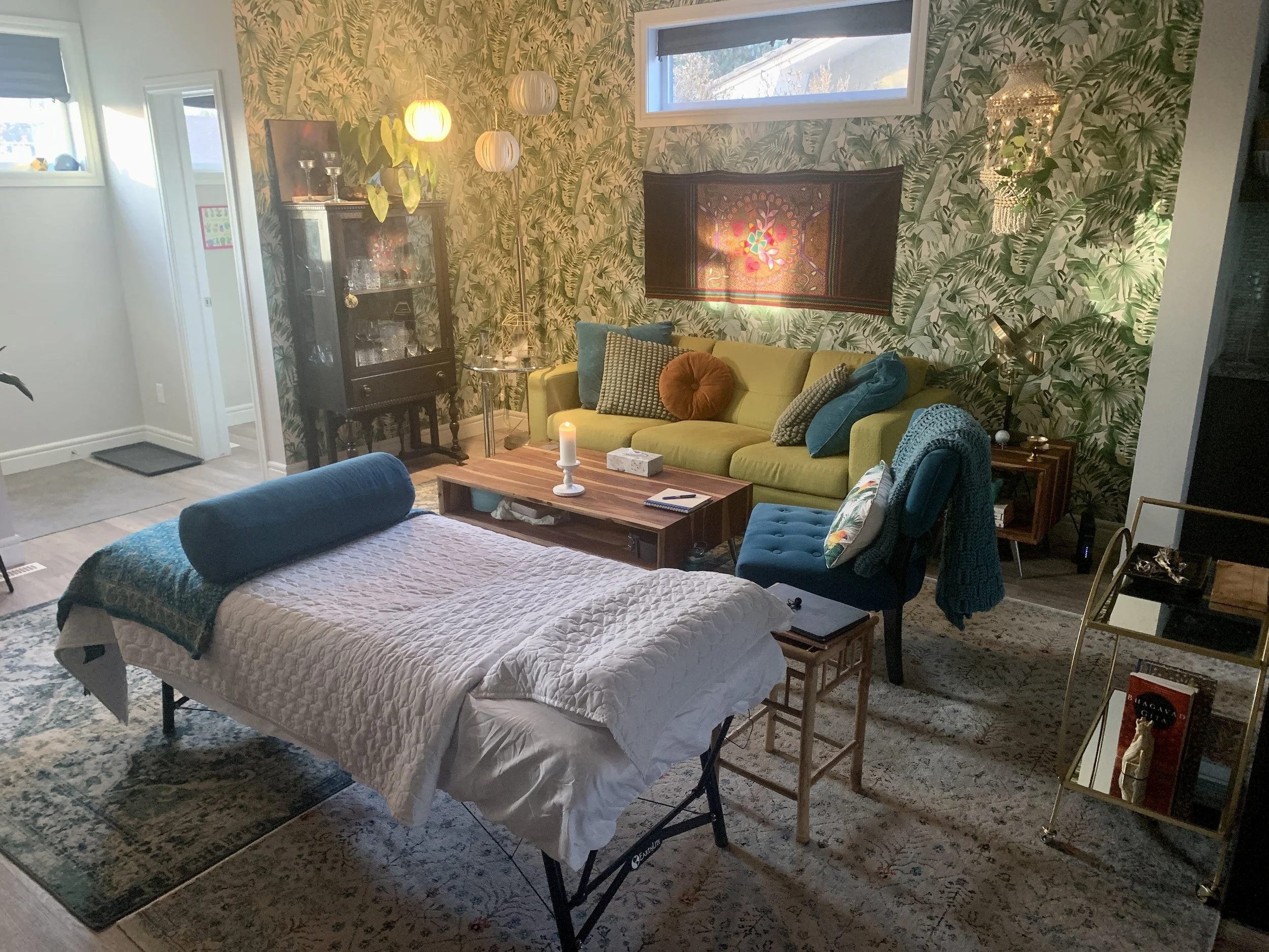 Living room with tropical wallpaper, yellow sofa, various pillows, art piece on wall, window, glass cabinet, small lamp, coffee table, massage table, blue chair with blanket, bookshelf, and decorative items.